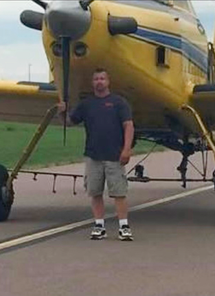 Man standing in front of a yellow helicopter on tarmac, wearing a dark shirt, khaki shorts, and sneakers.