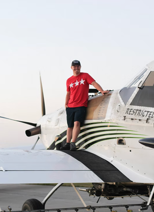 Man standing on top of a small private jet aircraft on the tarmac, wearing a red t-shirt with white stars, black shorts, and a baseball cap.