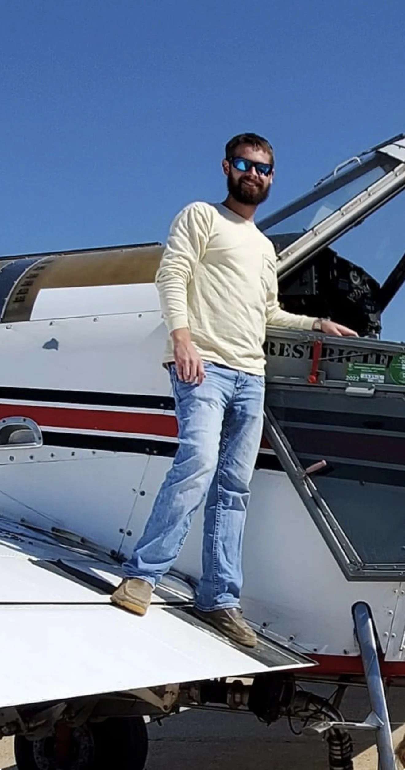 A man with sunglasses, a beige sweatshirt, and jeans standing on the wing of a small aircraft against a blue sky.