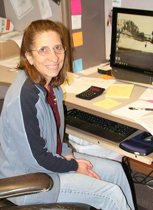 A woman sitting at a cluttered office desk with a computer monitor, keyboard, calculator, and various papers and sticky notes.