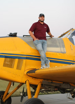 Man standing on the wing of a yellow airplane with blue stripe, wearing a maroon polo shirt and jeans, smiling at the camera.