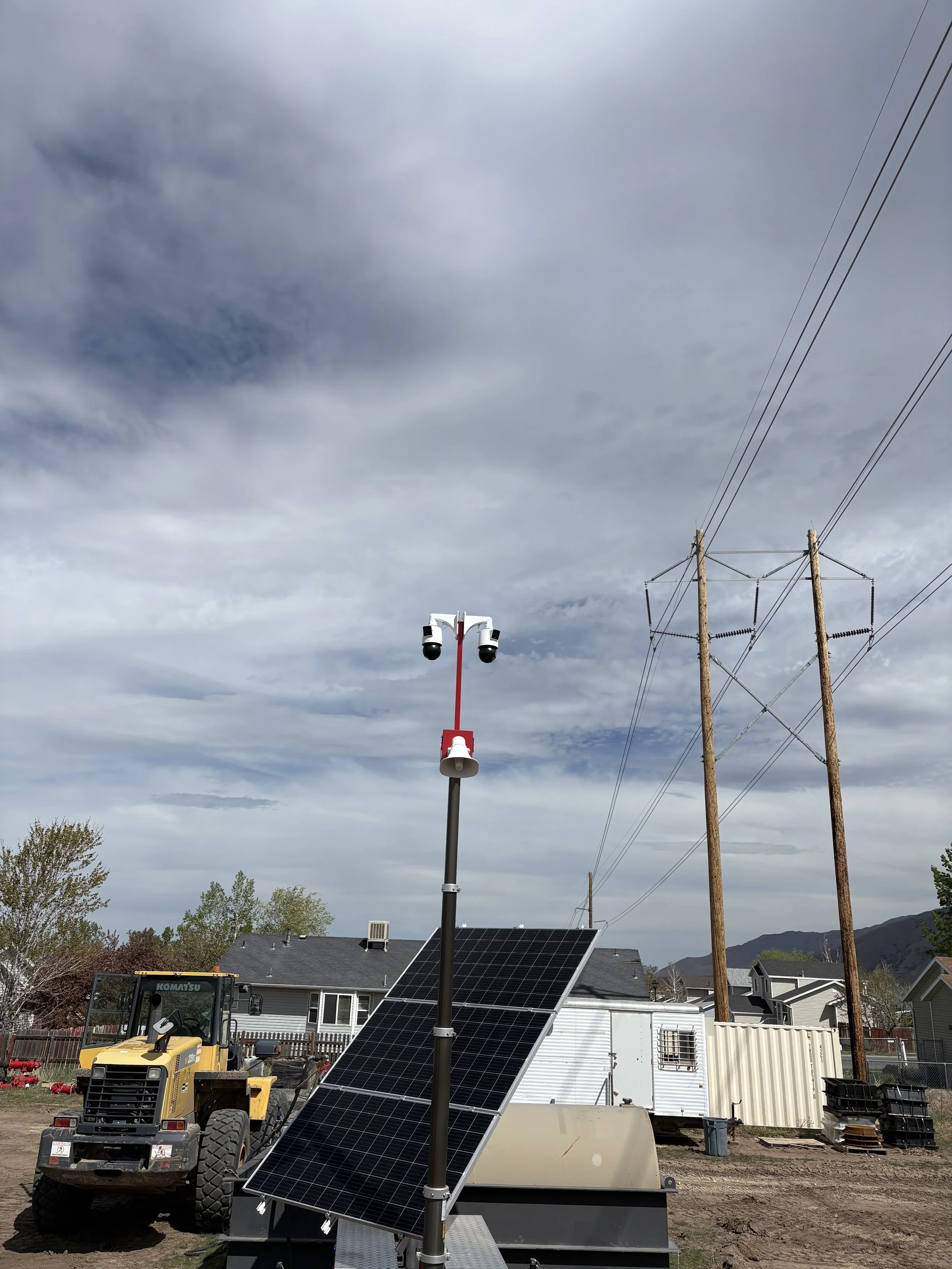 Solar panel installation with solar panels on a trailer, a tall pole with multiple security cameras, and utility poles against a cloudy sky in a residential area.
