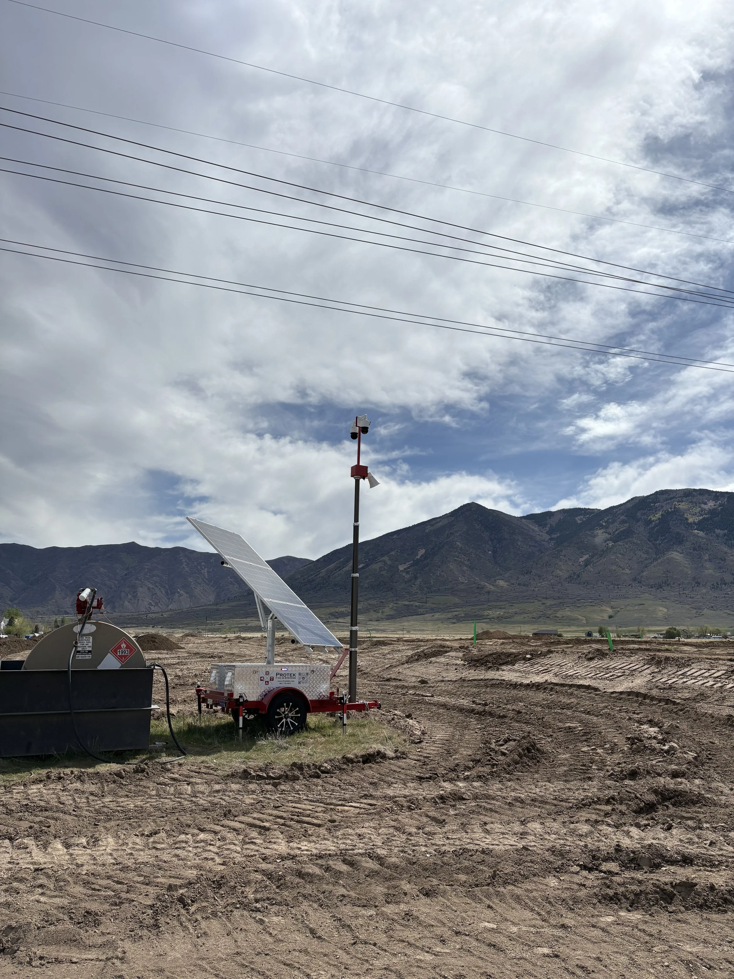 A solar-powered weather station with solar panels, a tall pole with sensors, and a large tank in a field with tracks. Mountains and cloudy sky in the background.