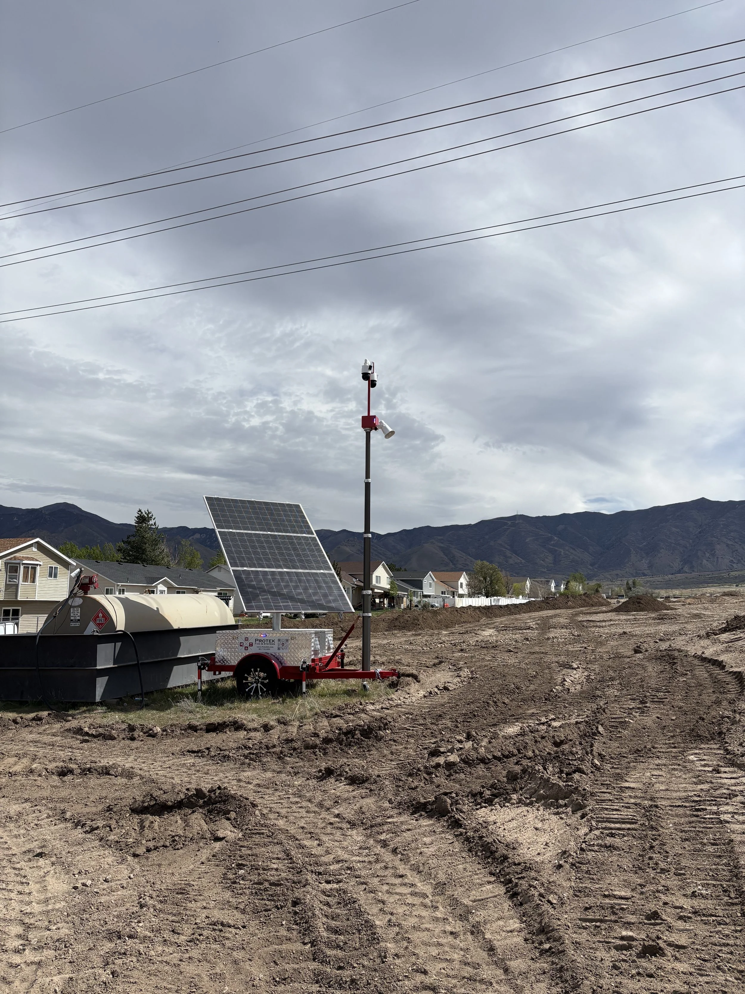 A solar-powered weather monitoring station with a tall pole, solar panels, and various sensors on a construction site with mountains in the background, cloudy sky, and nearby residential houses.