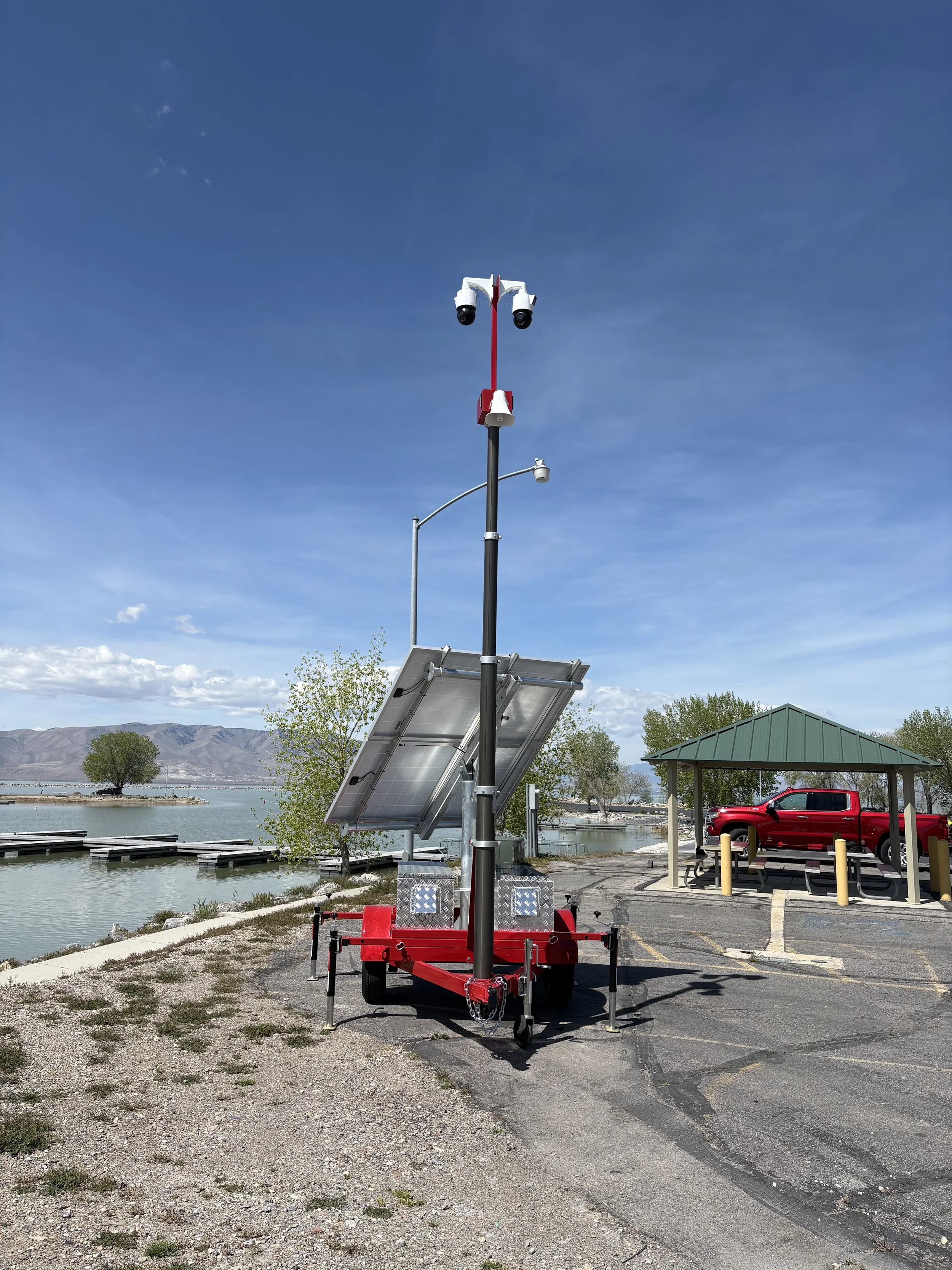 A mobile solar-powered traffic camera setup by a waterfront with a solar panel, a red truck parked under a shelter, and a vending machine nearby.