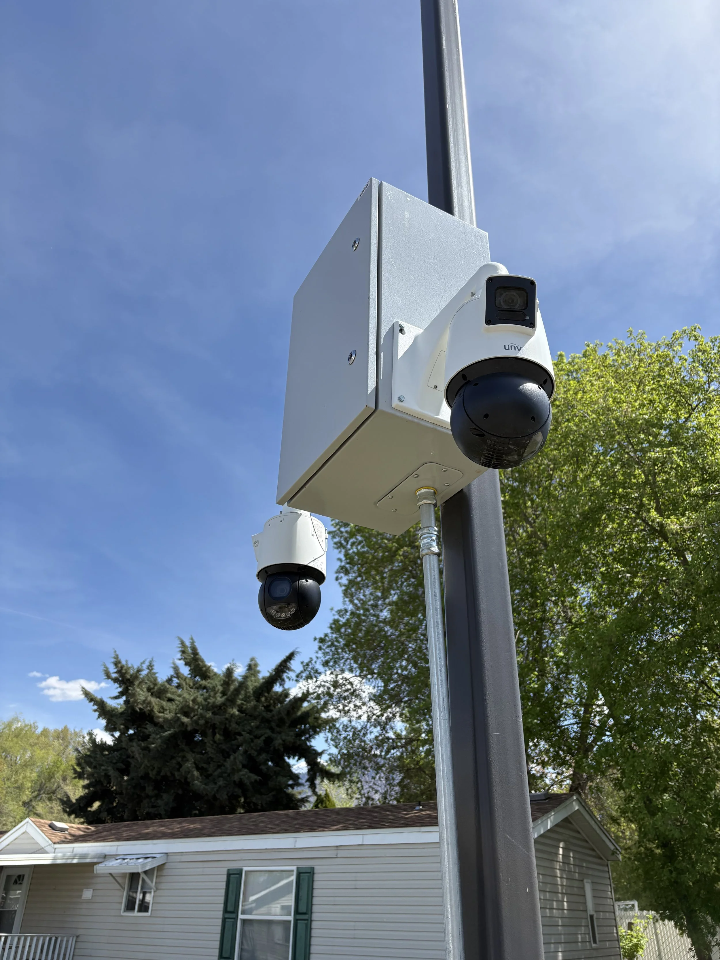 Close-up of a security camera mounted on a pole in front of a house with trees and a blue sky in the background.