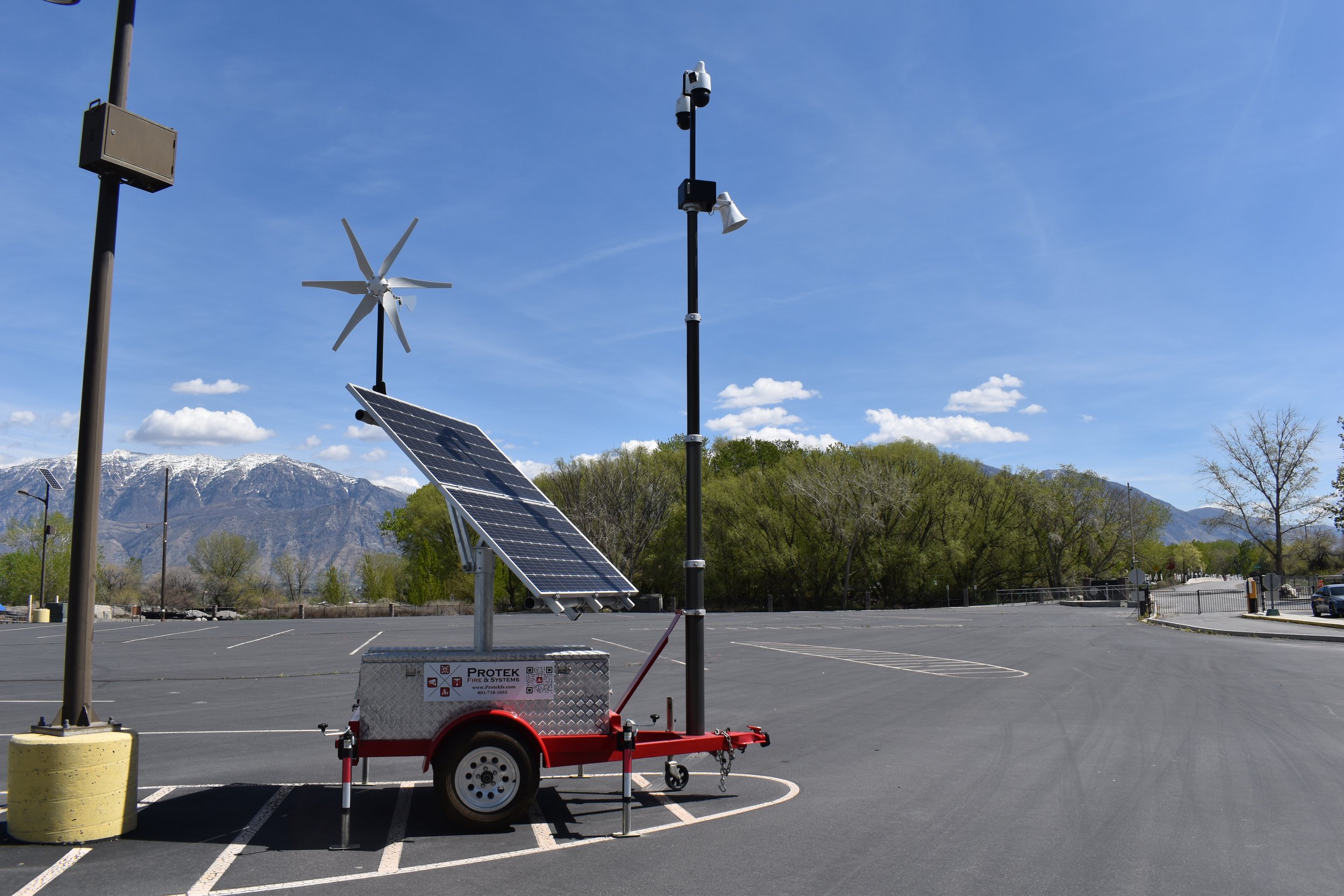 A mobile solar and weather monitoring station with solar panel, wind turbine, and sensors in a parking lot with mountains and trees in the background.