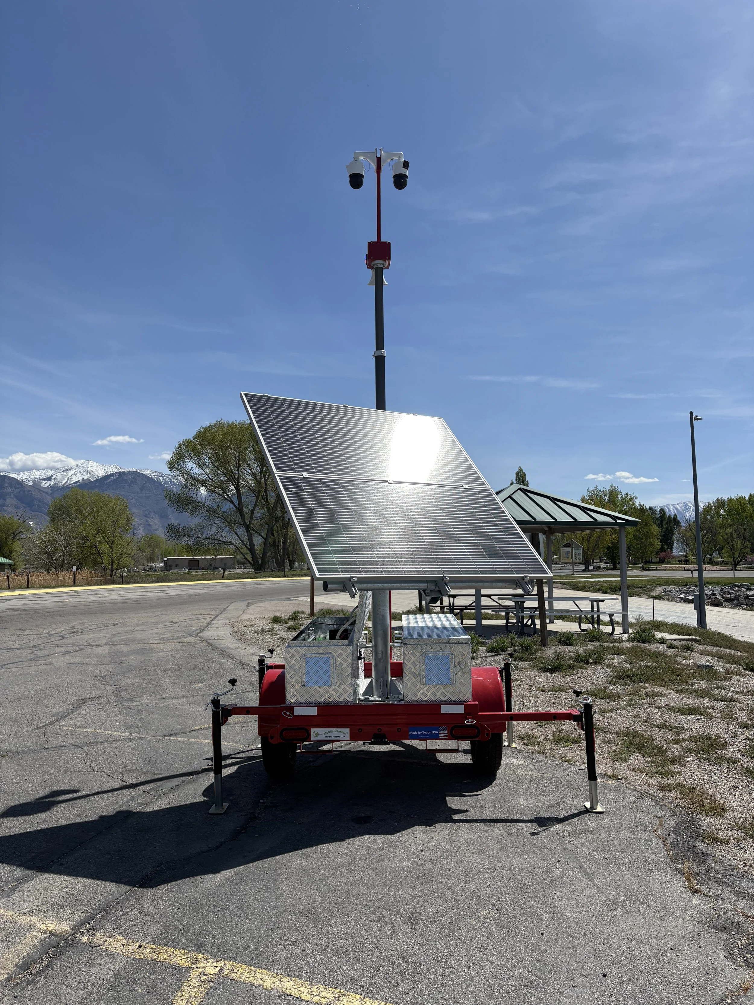 A mobile solar-powered surveillance camera system mounted on a trailer with solar panels, set up outdoors in a parking lot with mountains in the background.