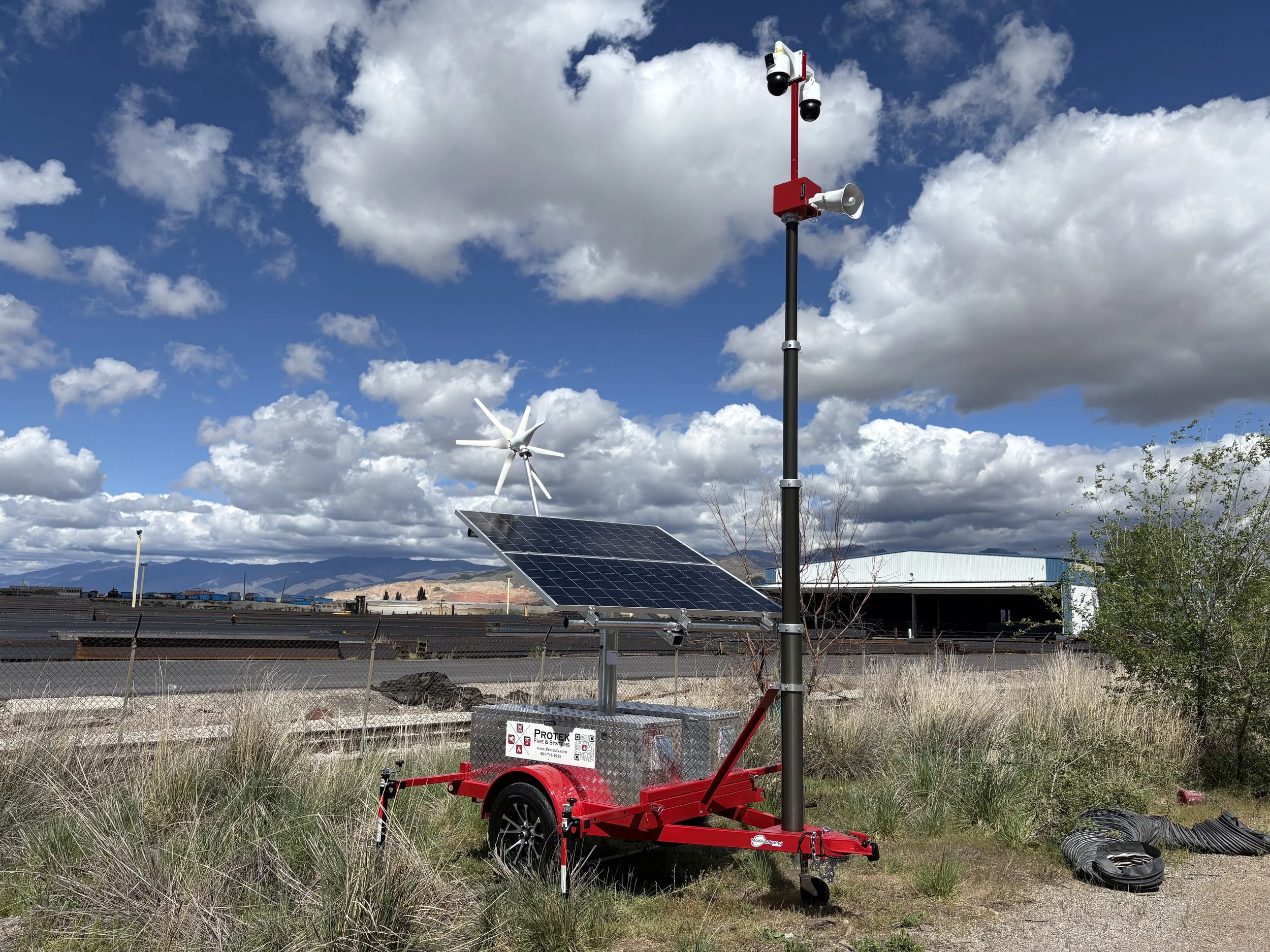 A mobile solar and wind power station with solar panels, a wind turbine, and surveillance cameras, set against a cloudy sky and mountains in the background.