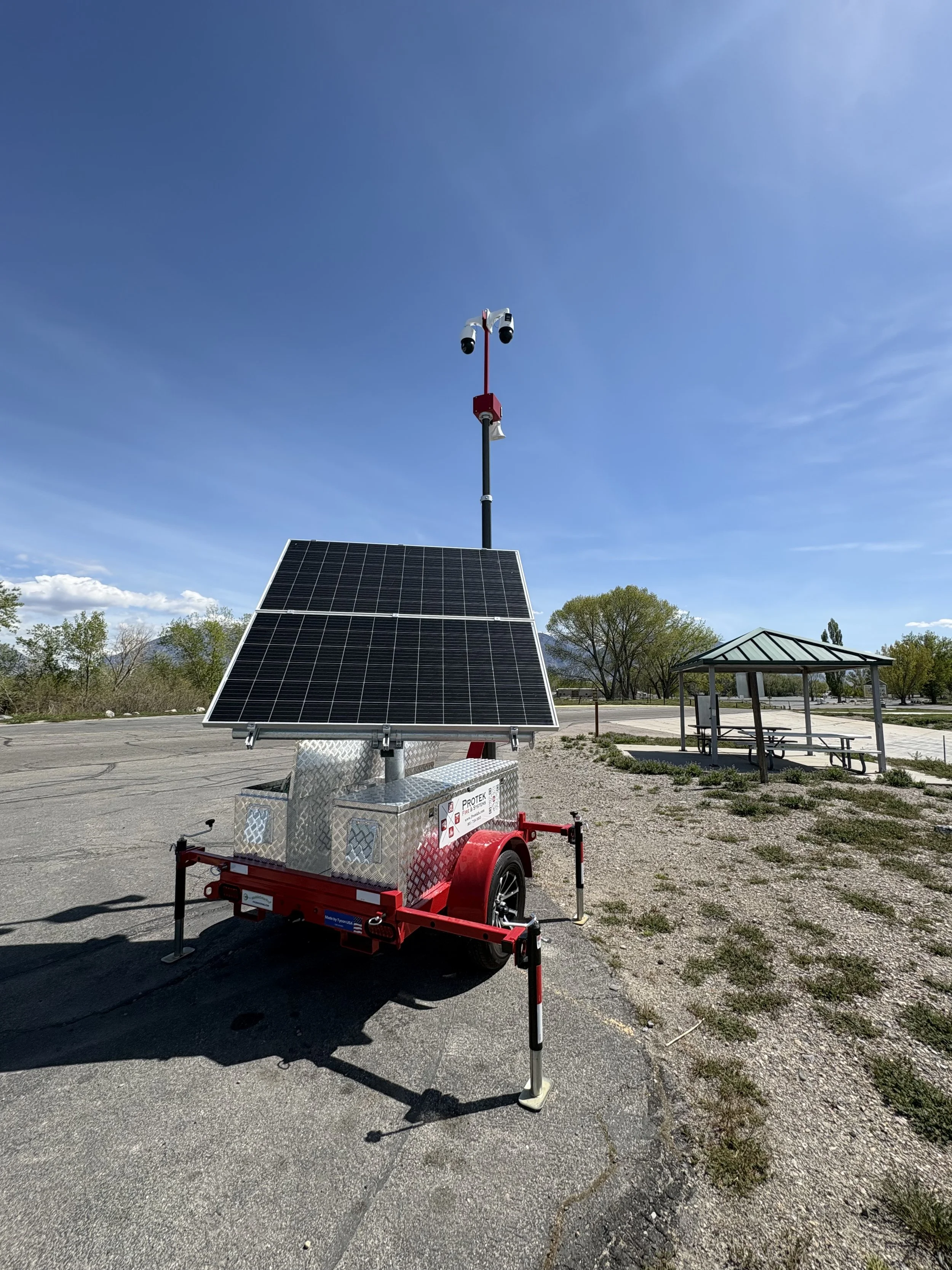 A trailer with a solar panel, two security cameras, and a weather alert system on a pole in an outdoor parking lot with trees and a blue sky.