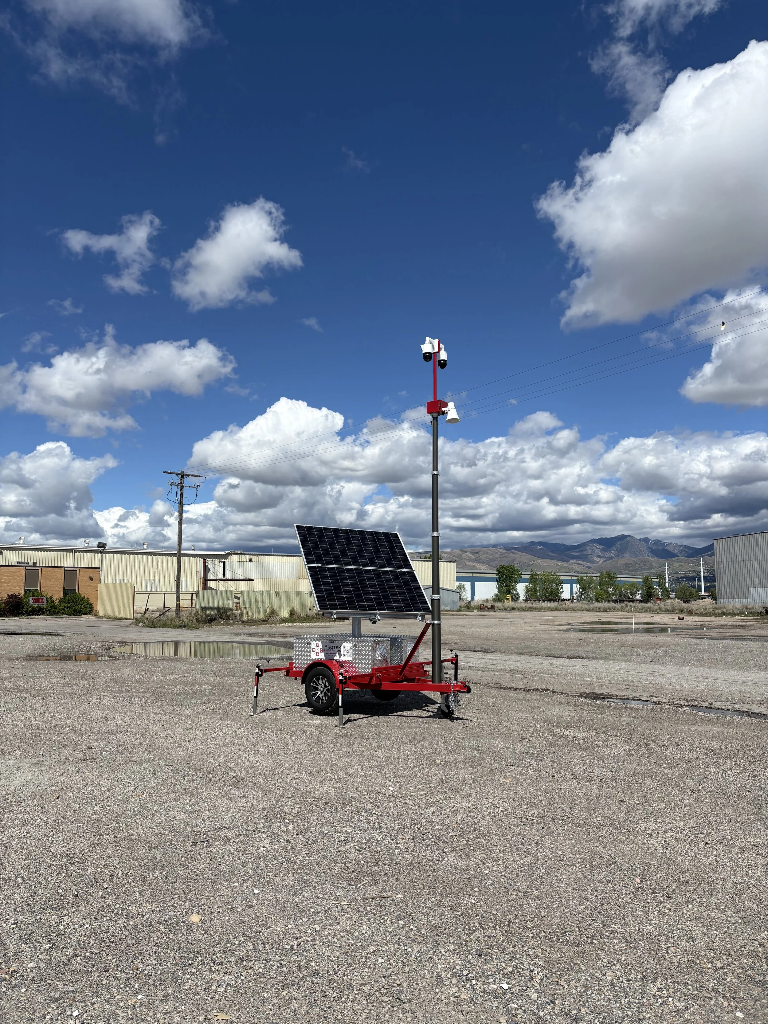 A weather monitoring station with solar panels, mounted on a red trailer, standing in an empty parking lot with industrial buildings and mountains in the background under a partly cloudy sky.
