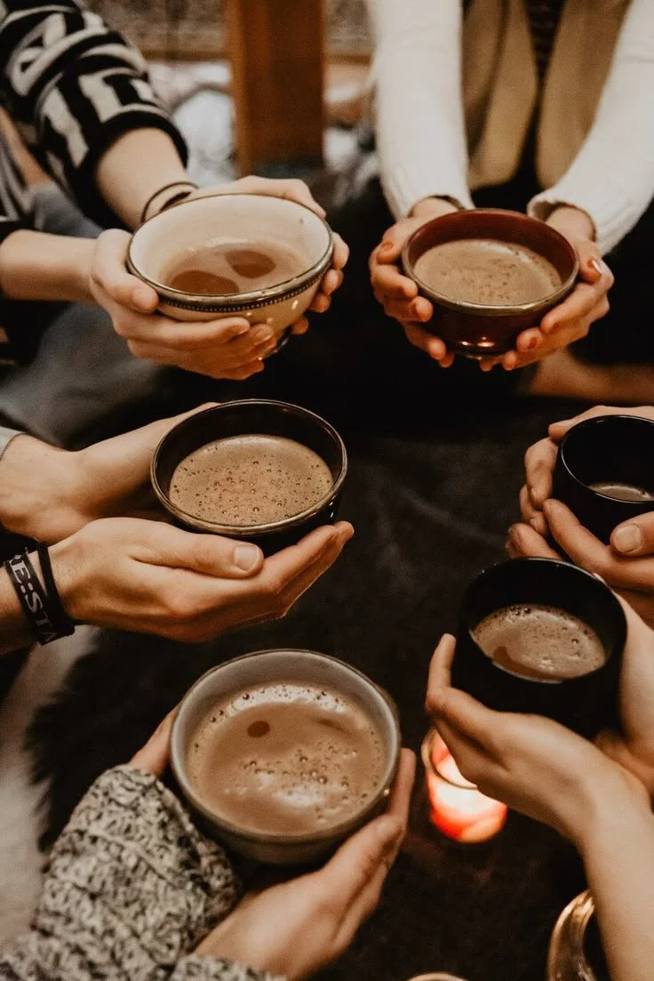 Multiple people holding cups of hot chocolate or coffee in a toast, with a candle on a dark table