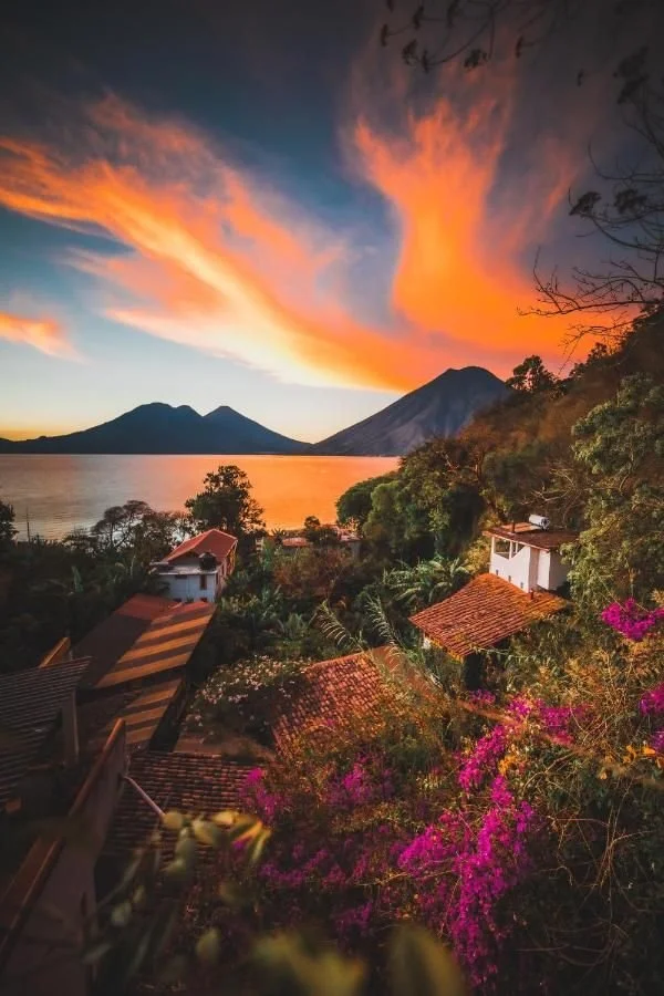 Sunset over a lake with mountains in the background, surrounded by lush trees and houses with reddish rooftops, and vibrant pink flowers in the foreground.