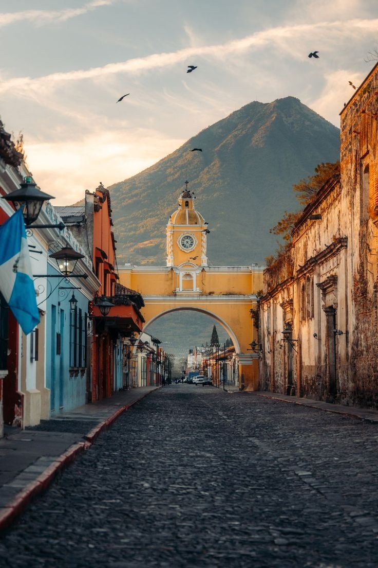 A cobblestone street leading to an archway with a yellow clock tower, colorful old buildings, and a mountain in the background. Birds are flying in the sky.