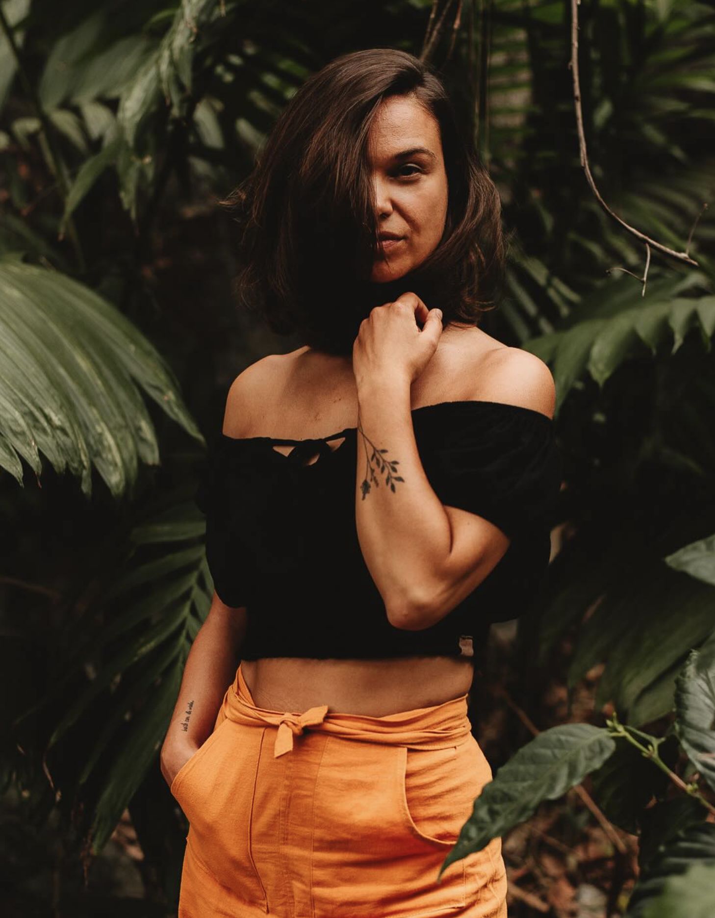 A woman standing amidst lush green tropical plants, wearing an off-the-shoulder black top and orange pants, with her hand near her face and a tattoo on her arm.