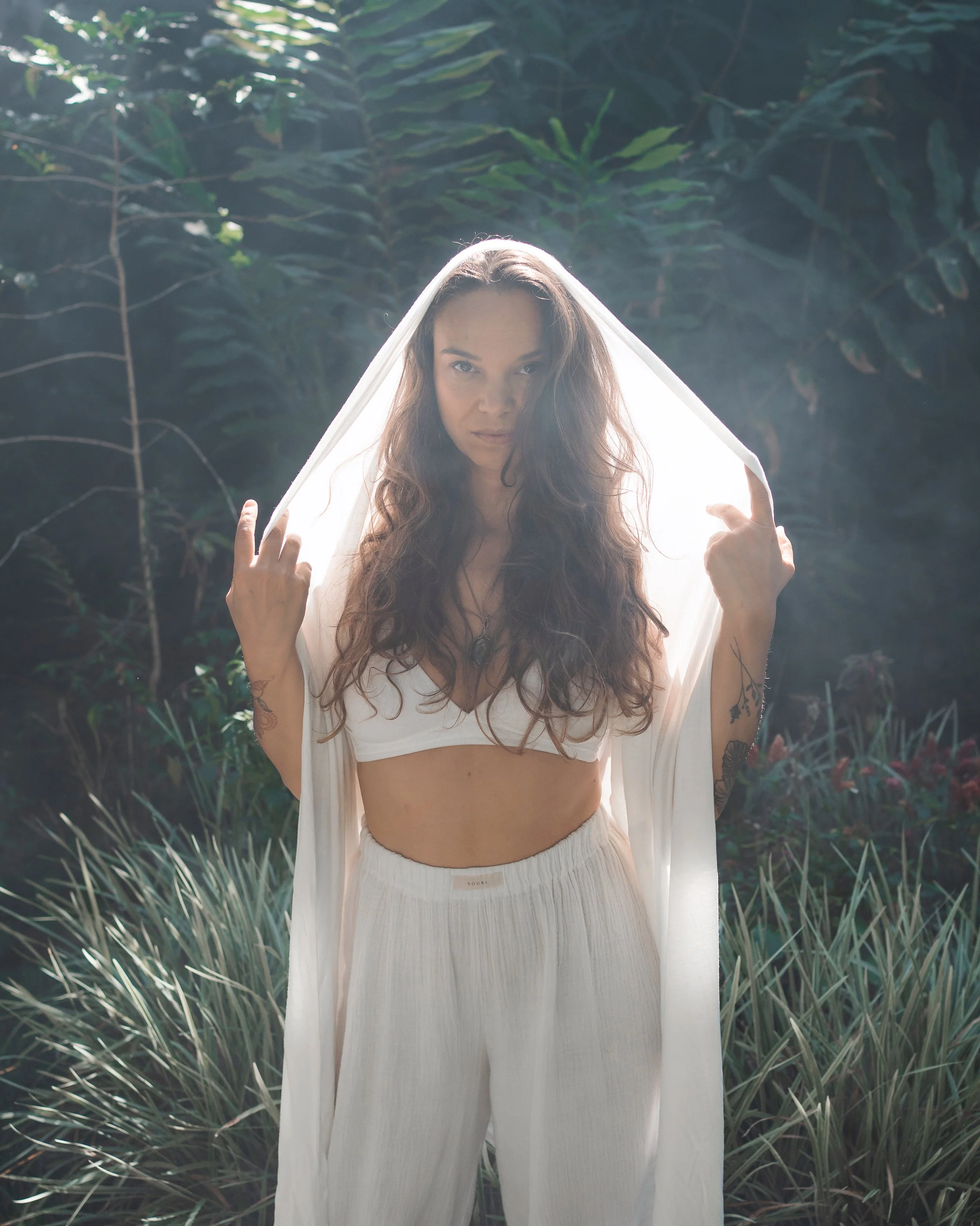 A woman with long, wavy brown hair standing outdoors among lush green plants and trees, holding a light-colored cloth or shawl over her head.
