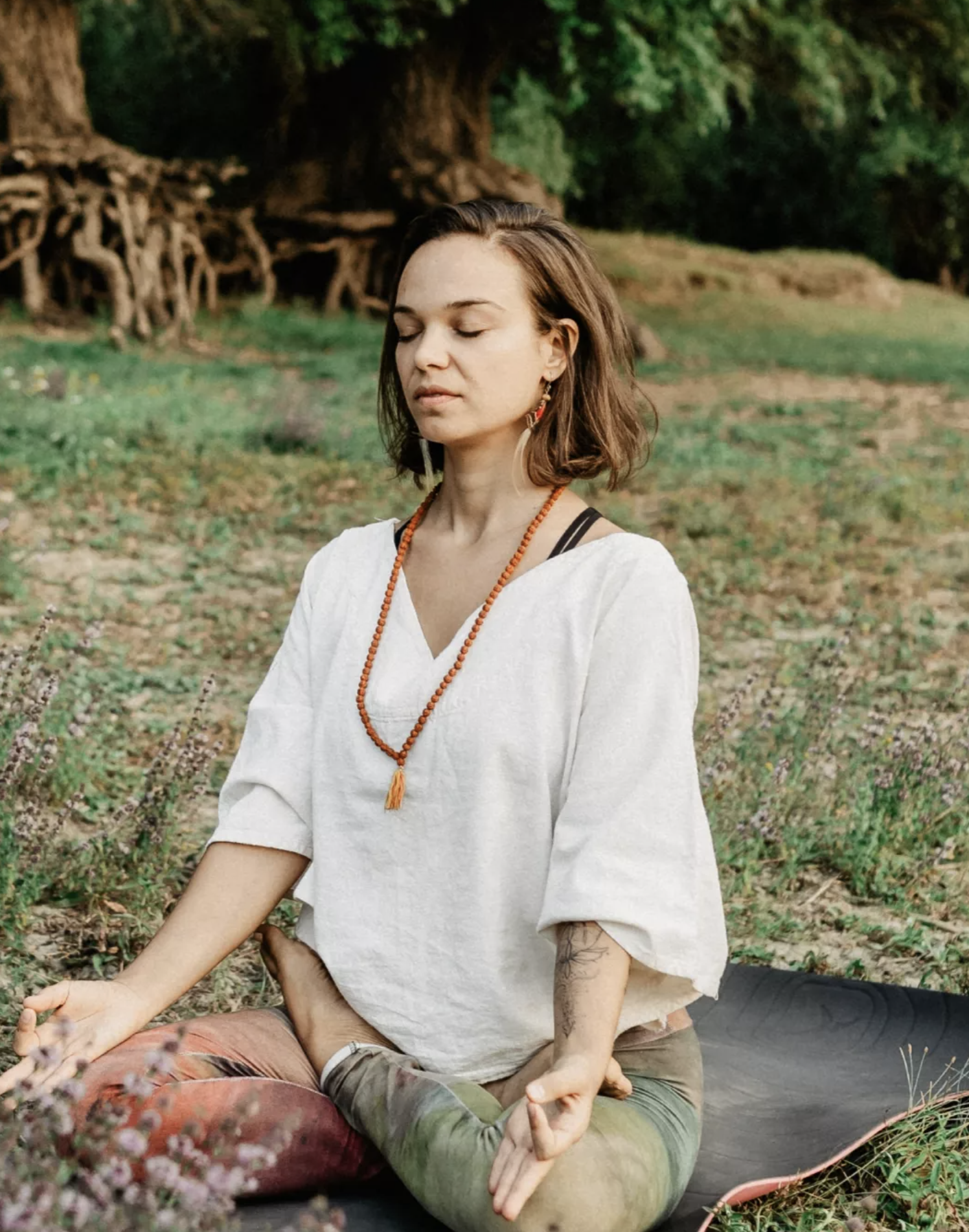 A woman practicing yoga outdoors in a nature setting, sitting on a yoga mat in a meditative pose with eyes closed, wearing a white top, beaded necklace, and colorful leggings.
