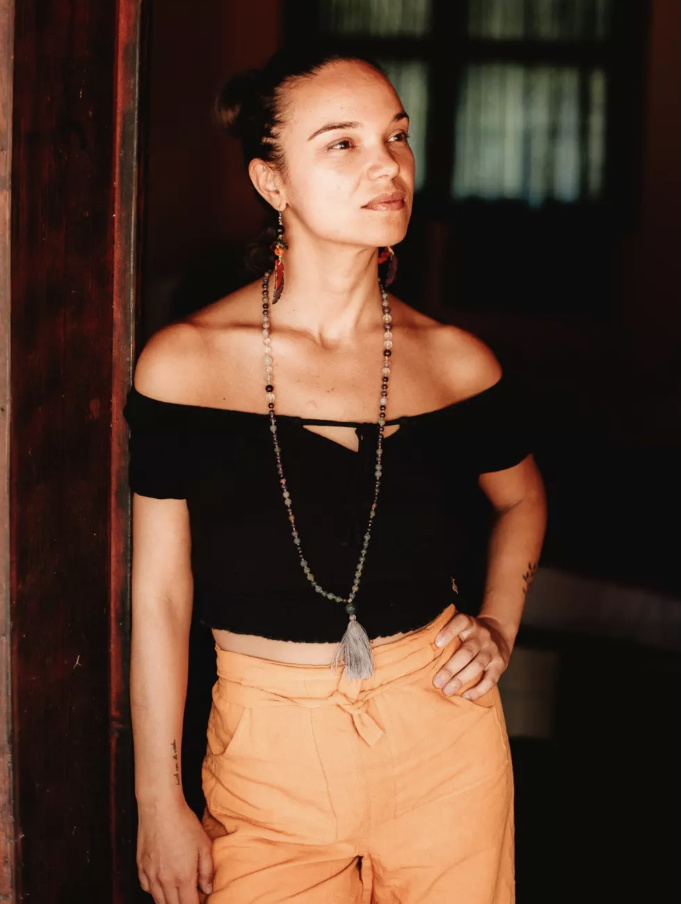 A woman with dark hair in a bun, wearing an off-the-shoulder black top and orange pants, standing indoors with a dark background and a window with curtains.