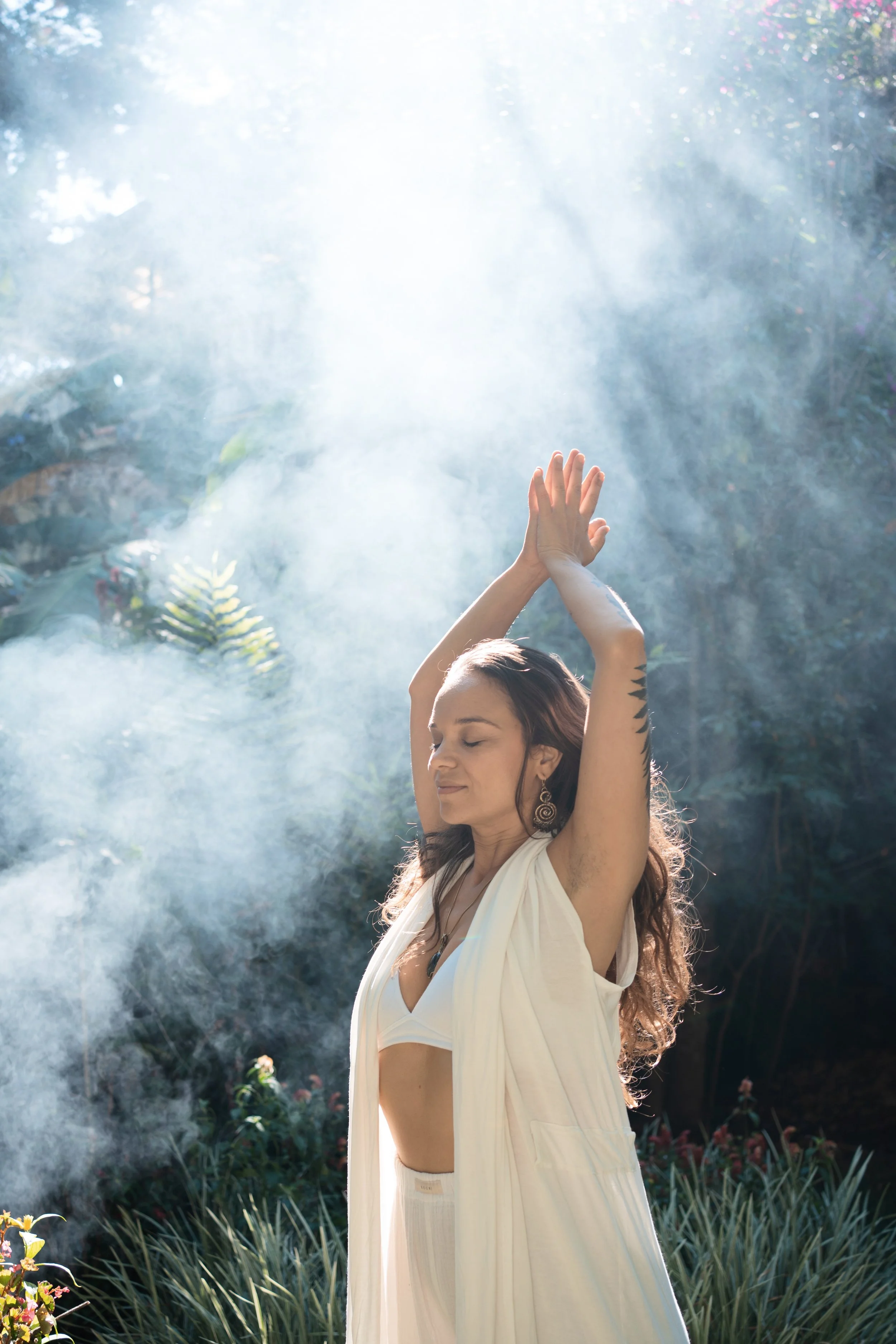A woman with long brown hair, wearing a white sleeveless top and loose white pants, stands outdoors with her eyes closed, with her arms raised above her head, clasping her hands, in a peaceful pose. She is surrounded by greenery and mist.