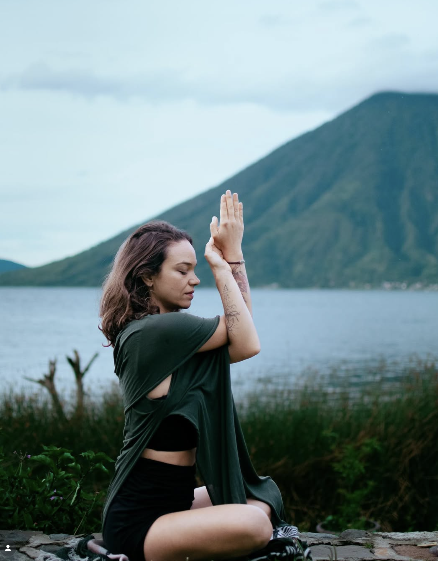Young woman practicing yoga outdoors by a lake with mountains in the background, sitting cross-legged with hands in prayer position and eyes closed.