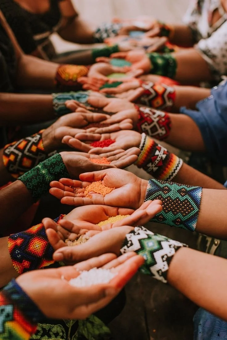 Multiple hands with colorful beaded bracelets reaching towards the camera, each hand holding small piles of different colored grains or powders.
