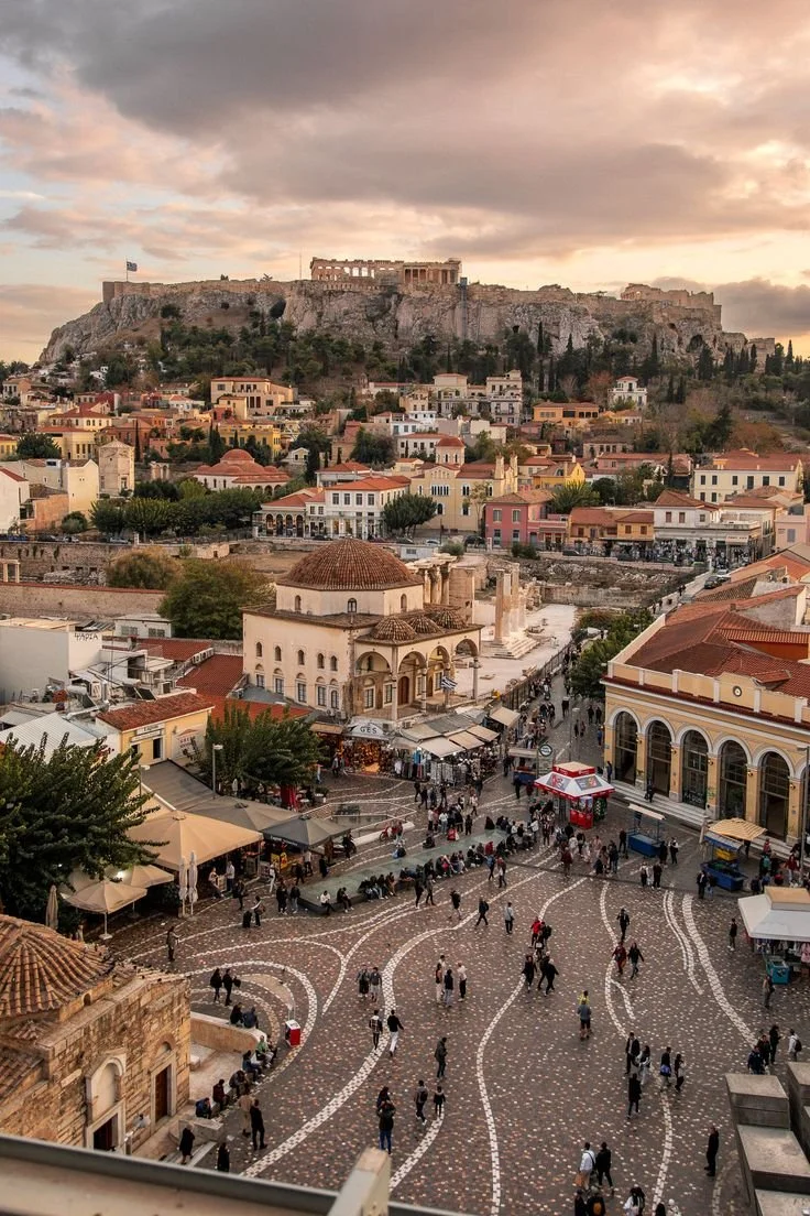 A historic city square with cobblestone streets filled with people, surrounded by colorful buildings, with the Acropolis and Parthenon on a rocky hill in the background at sunset.