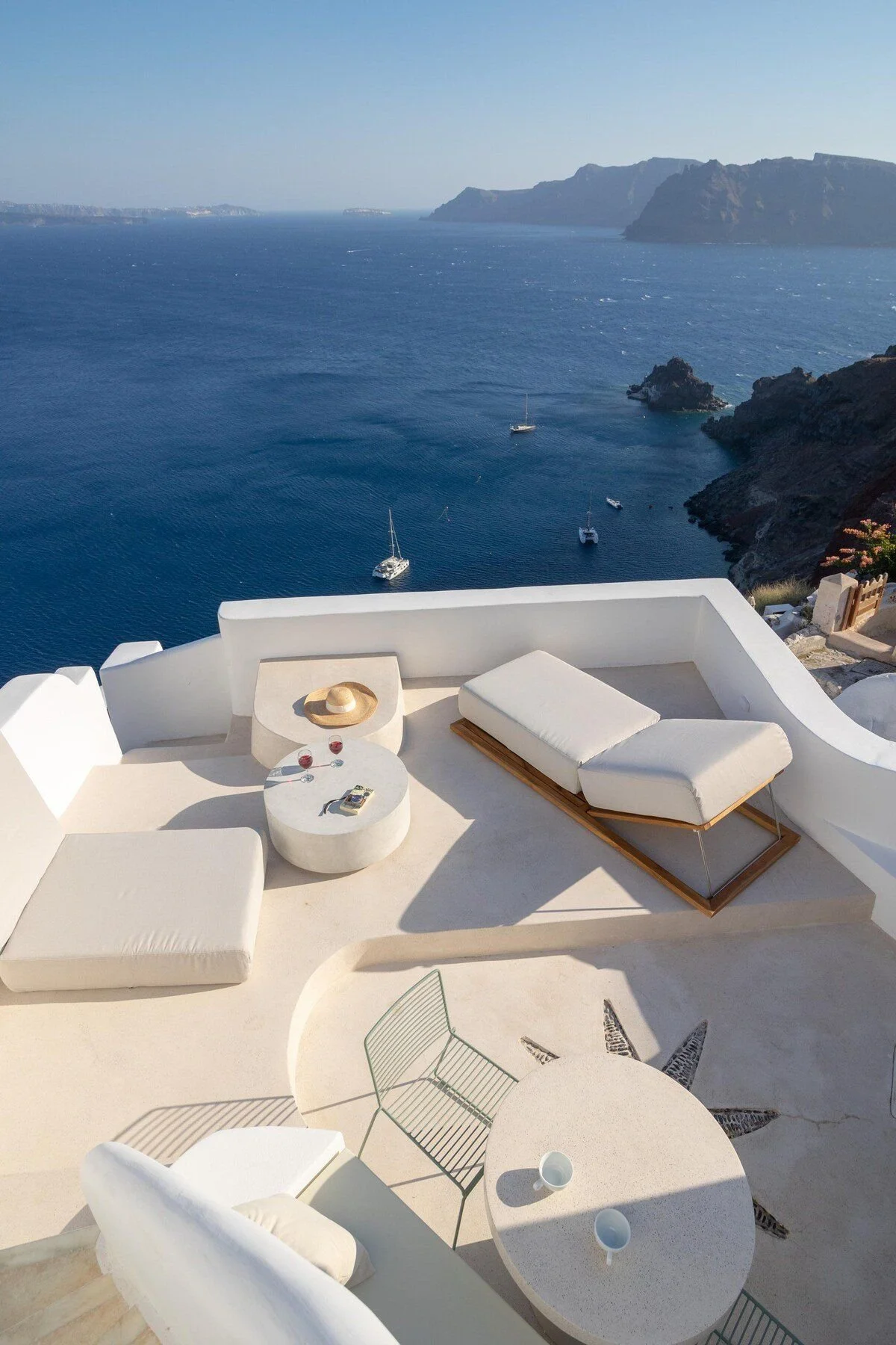 A white terrace overlooking a deep blue sea with boats and cliffs in the background, featuring outdoor furniture and minimalist decor.