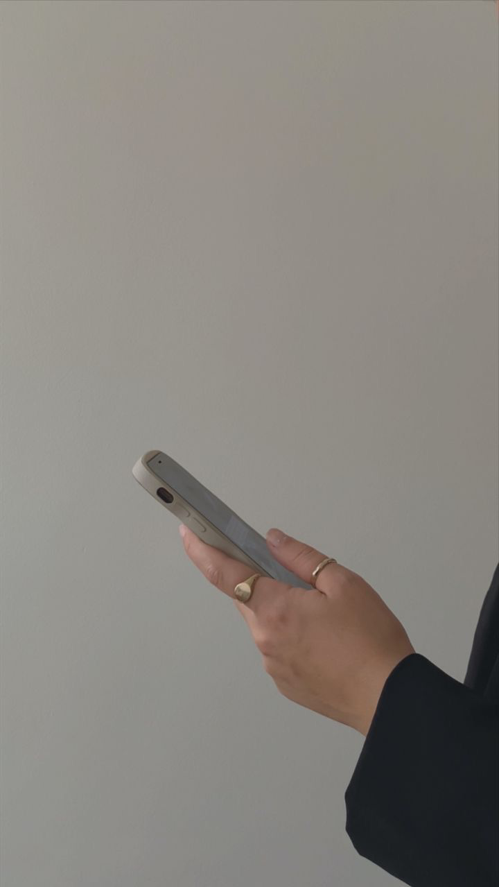 Person holding a smartphone in their right hand, with a light-colored wall in the background.
