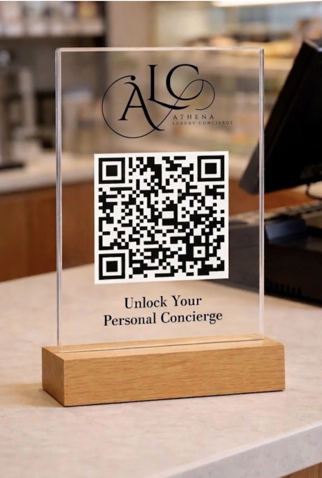 A clear acrylic sign with the logo 'A.L.C Athena Luxury Concierge' at the top, a QR code in the center, and the text 'Unlock Your Personal Concierge' at the bottom, placed on a wooden stand on a countertop.