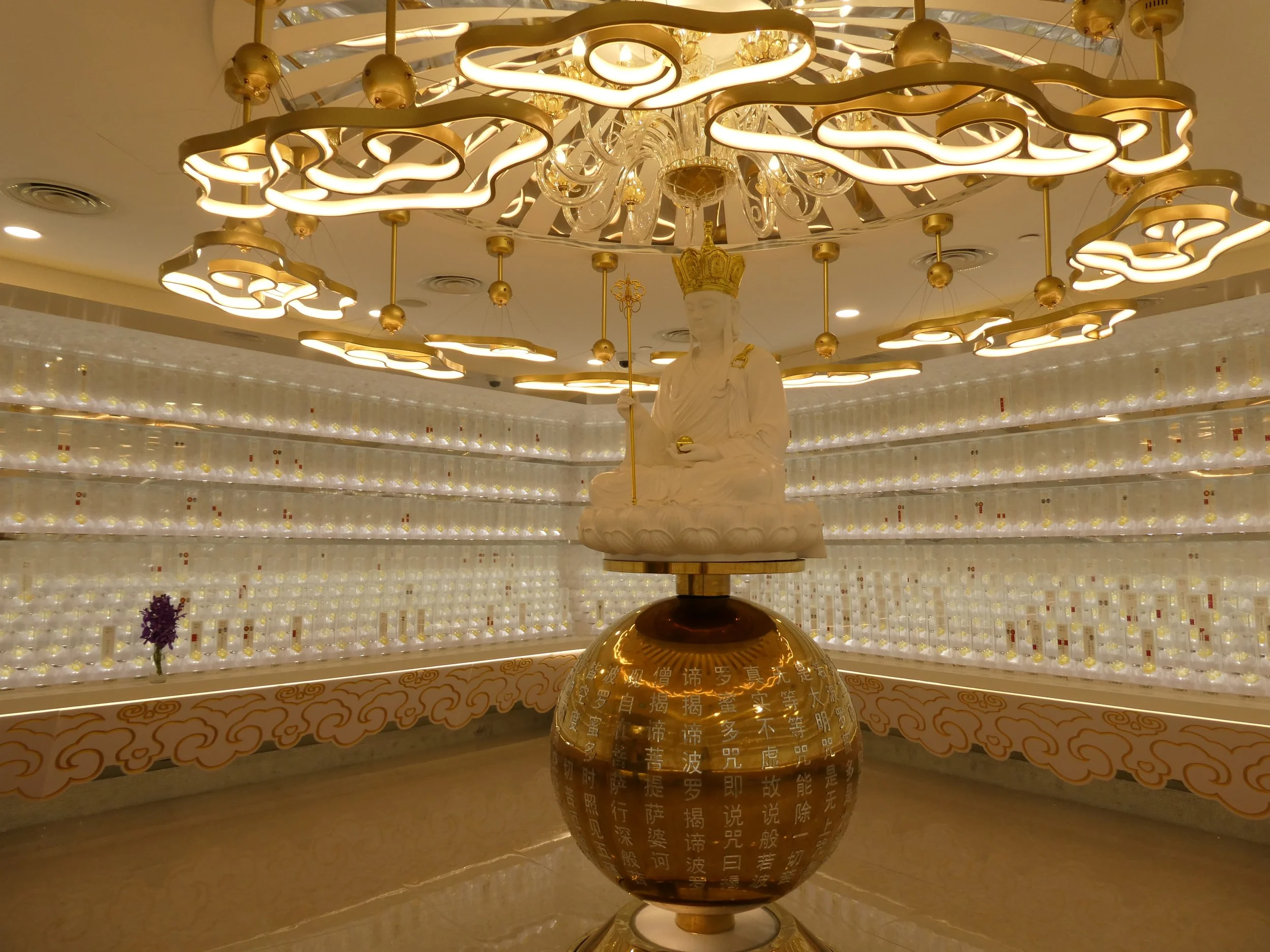 An ornate chandelier with gold accents and intricate neon light design, hanging above a statue of a seated figure wearing a crown, surrounded by rows of small illuminated bottles with Chinese characters on a wall behind.