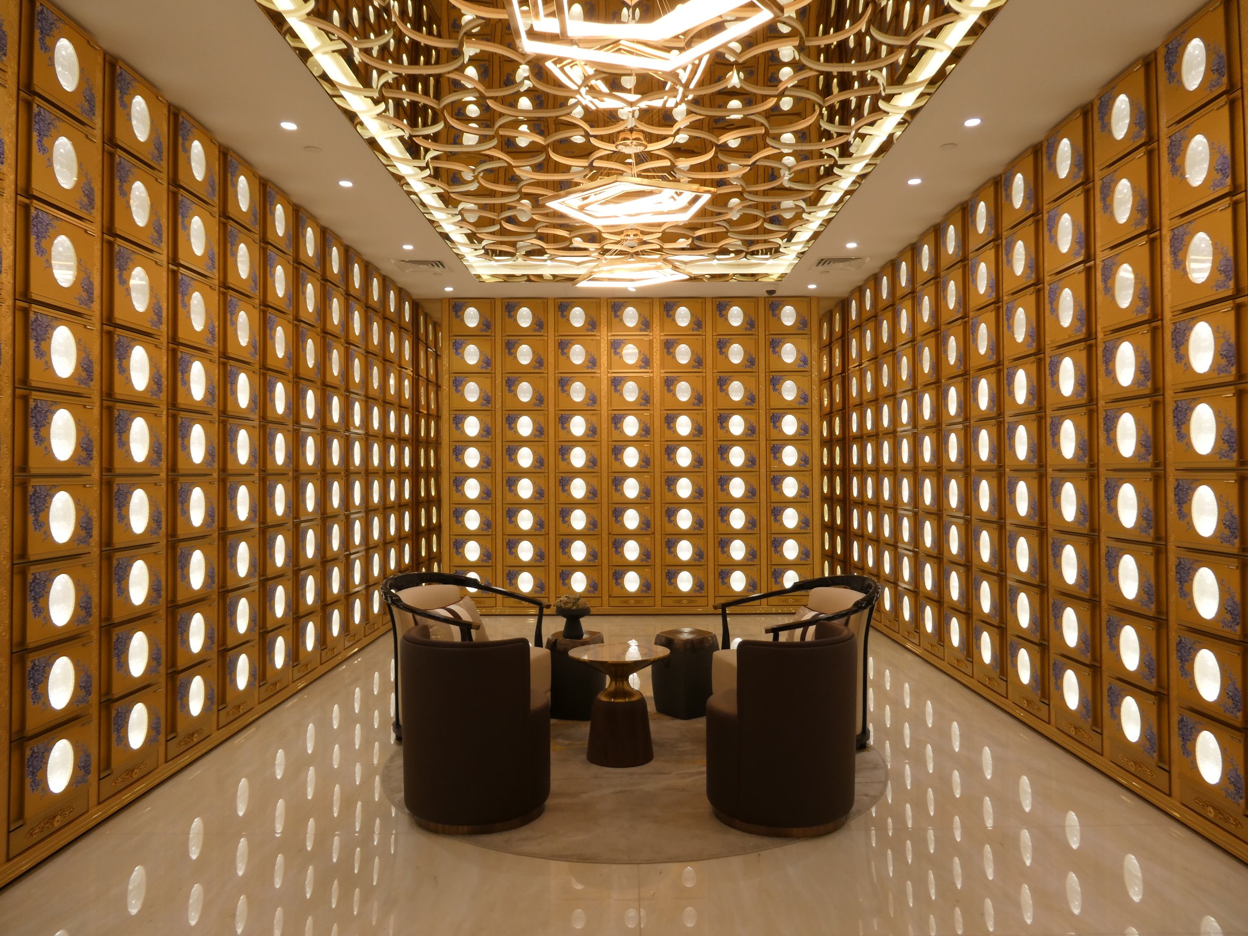 Interior of a luxurious room with gold-colored walls featuring oval mirrors, a modern ceiling light fixture, and a seating area with black chairs and a small round table.