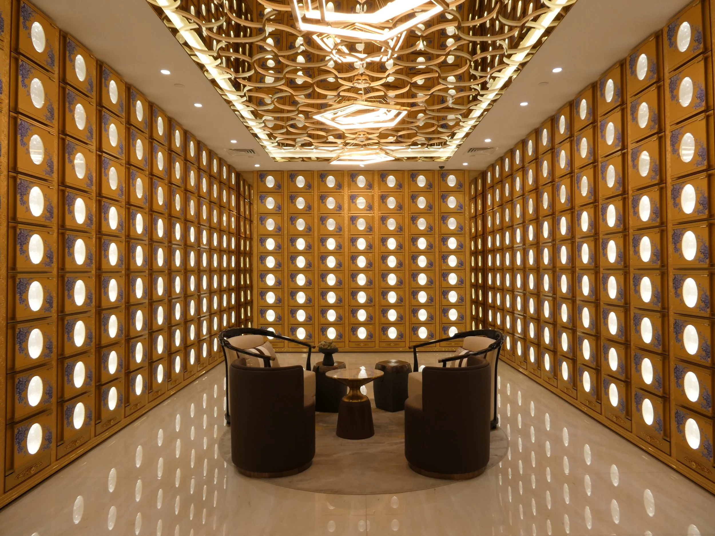 A modern columbarium suite with walls covered in wooden boxes with circular cutouts, a geometric light fixture on the ceiling, and a seating area with four chairs and a small round table in the center.