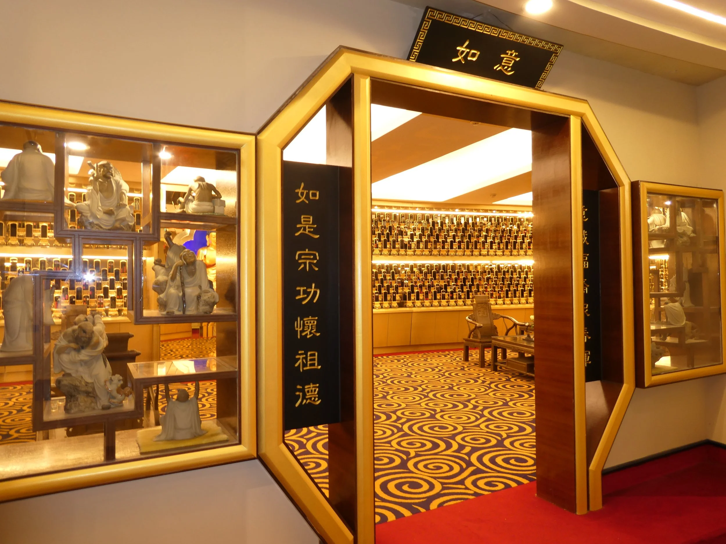 Decorative display with Chinese characters and sculptures of figures, including a seated man and a person praying, set against a background of illuminated shelves.