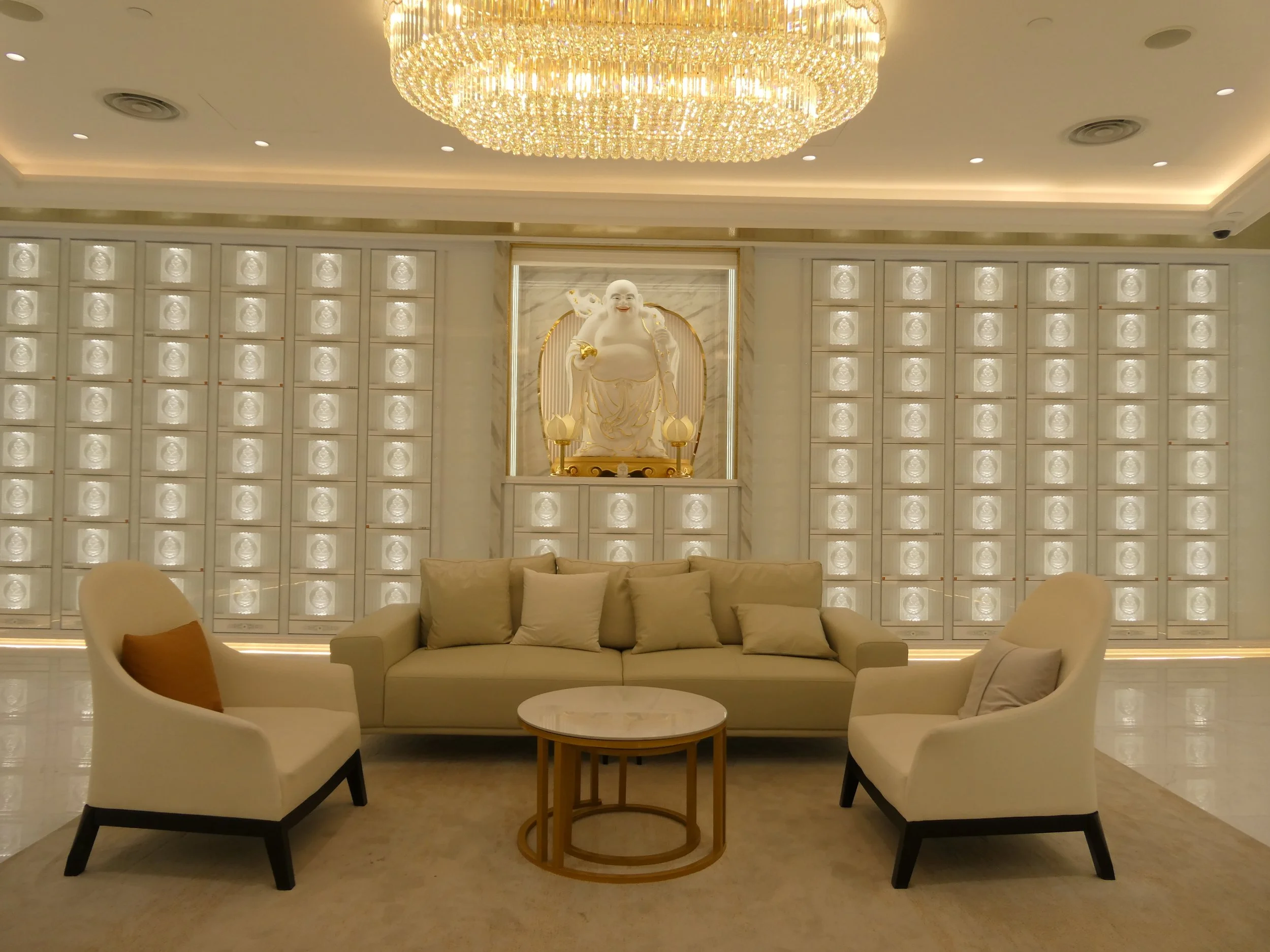 Elegant columbarium suite with white walls, a large crystal chandelier, a Buddha statue on a marble wall, and white sofas with cushions.