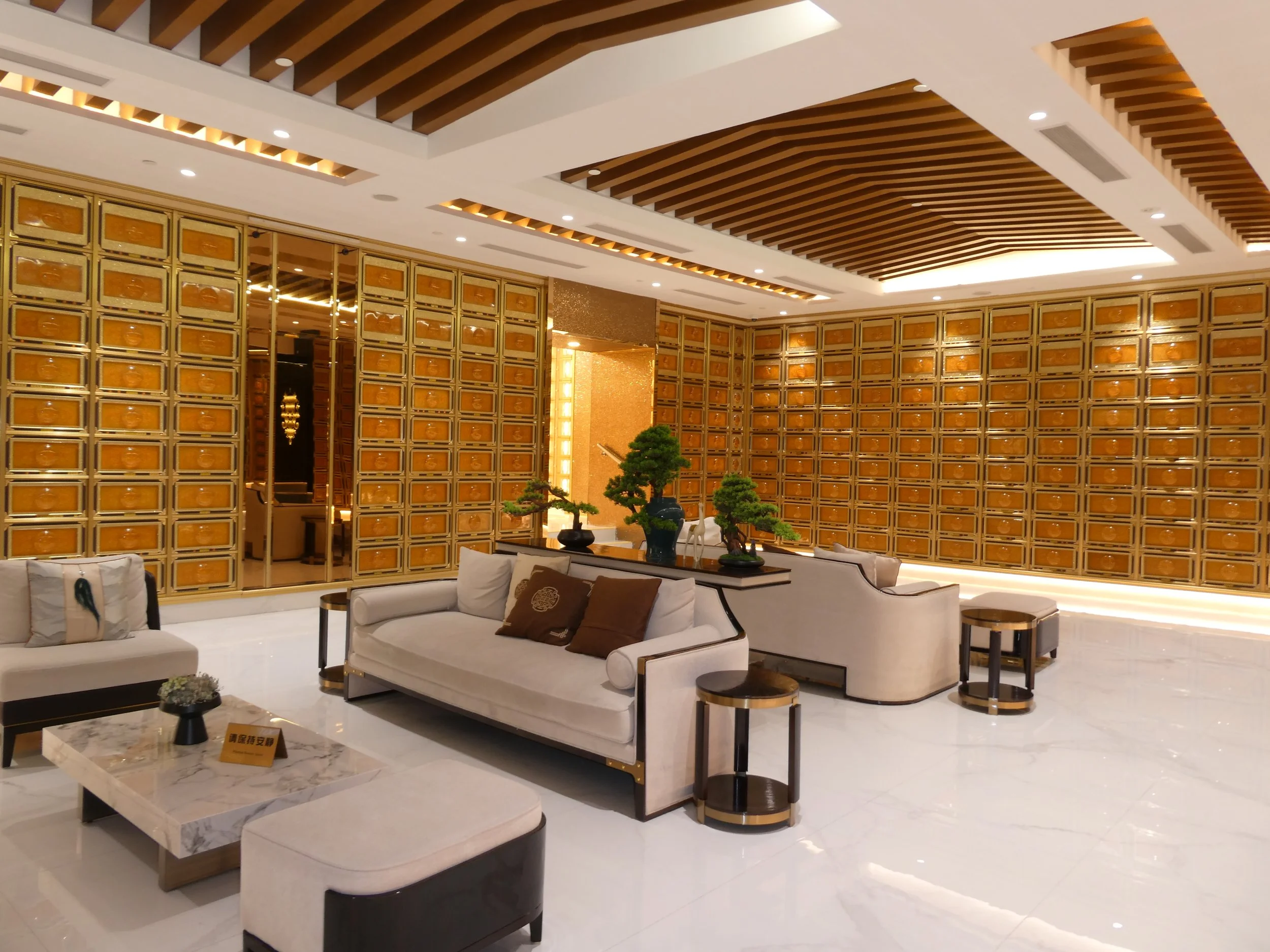 Luxurious hotel lobby with white and gold furniture, decorative plants, and a ceiling with wooden slats and embedded lighting.