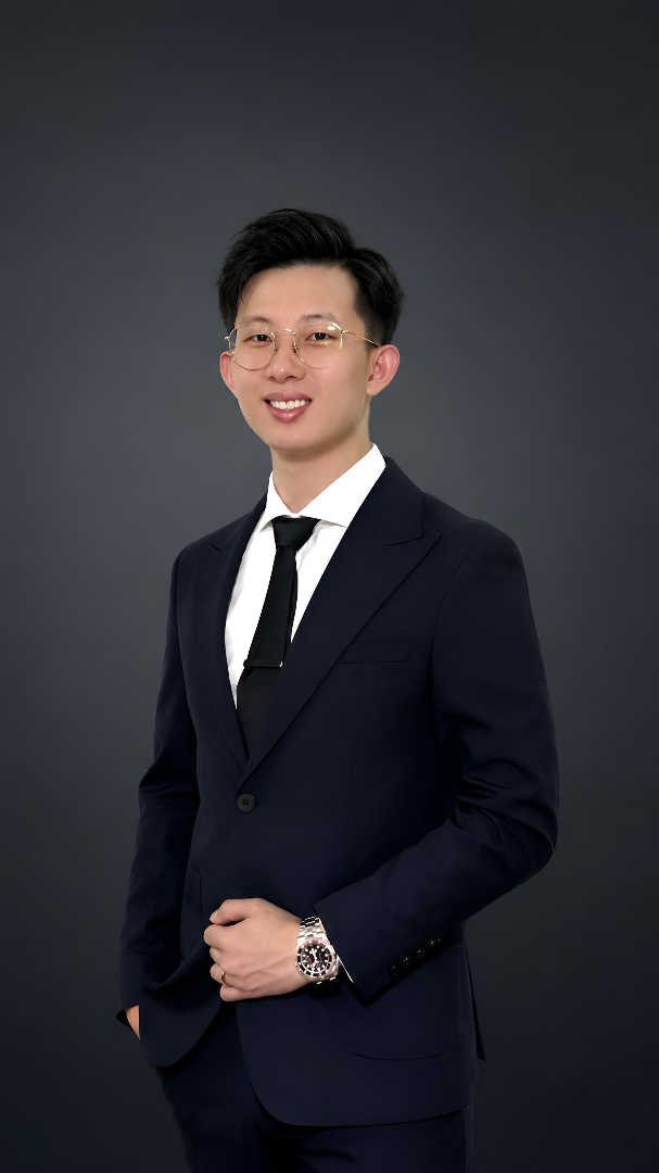 A young man wearing glasses in a dark suit, white shirt, and black tie, standing against a dark gray background with a slight smile and his left hand resting on his belt.