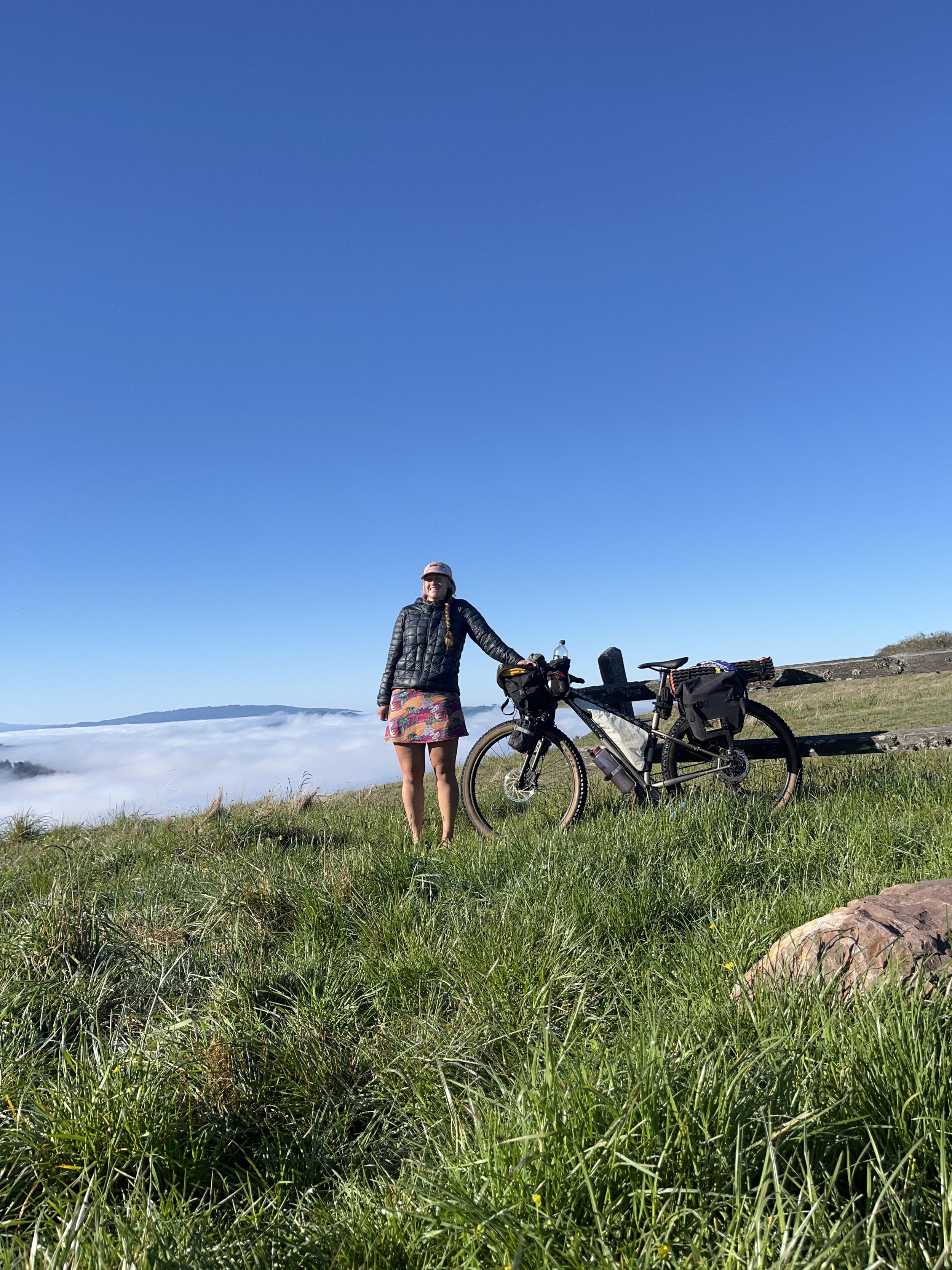 A woman stands on a grassy hill with a bicycle beside her, overlooking a cloud-covered landscape under a clear blue sky.