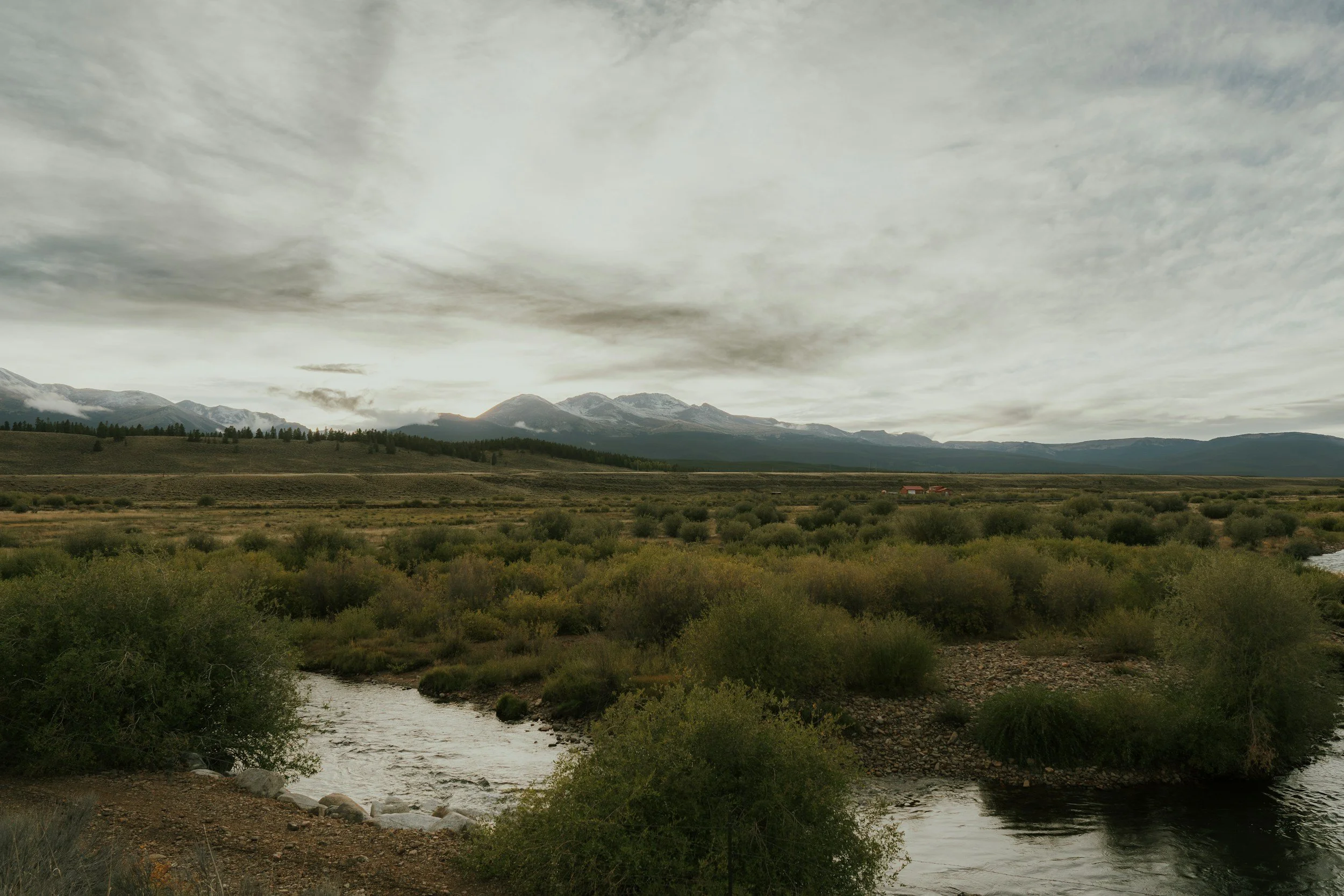 A landscape with a river in the foreground, rolling green hills, distant mountains, and a cloudy sky.