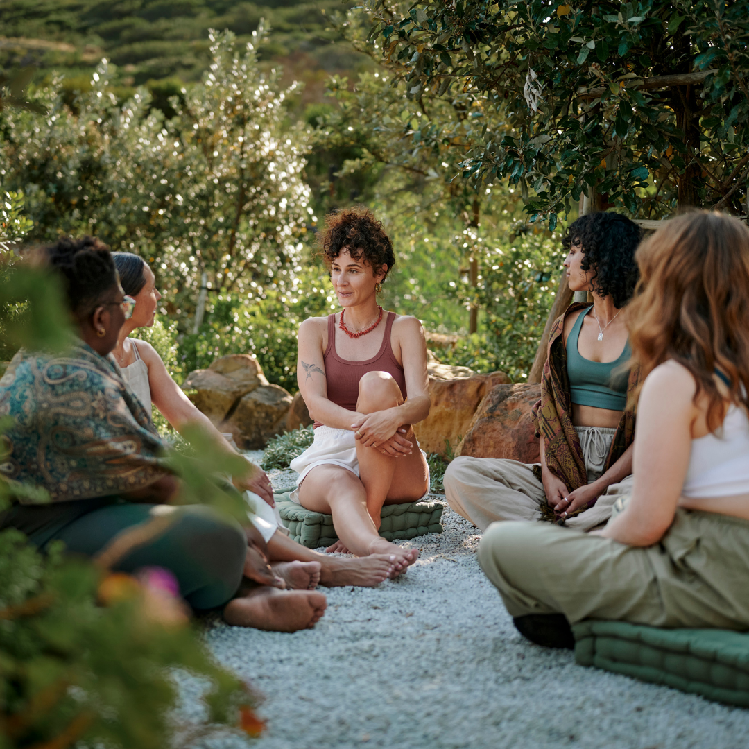 Group of five women sitting outdoors on cushions, engaged in conversation in a lush garden setting.