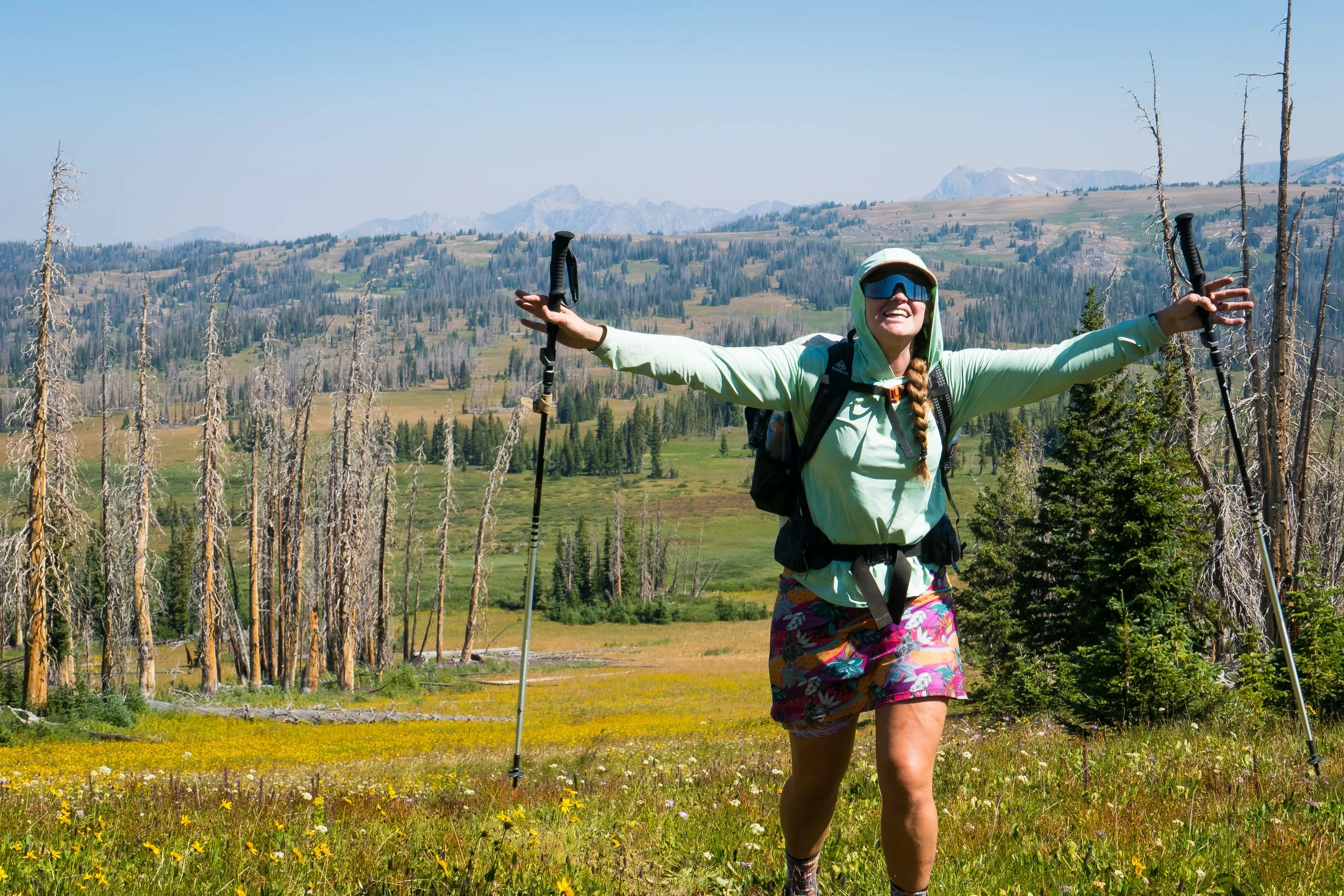 A woman hiking in a mountainous landscape with open arms and a big smile, holding trekking poles, wearing a green jacket, colorful shorts, and sunglasses.