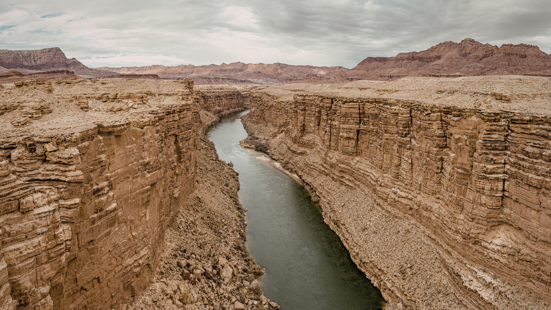 A canyon with tall, layered rock walls on either side of a narrow river flowing through the bottom, with rugged desert terrain and distant mountains under cloudy skies.