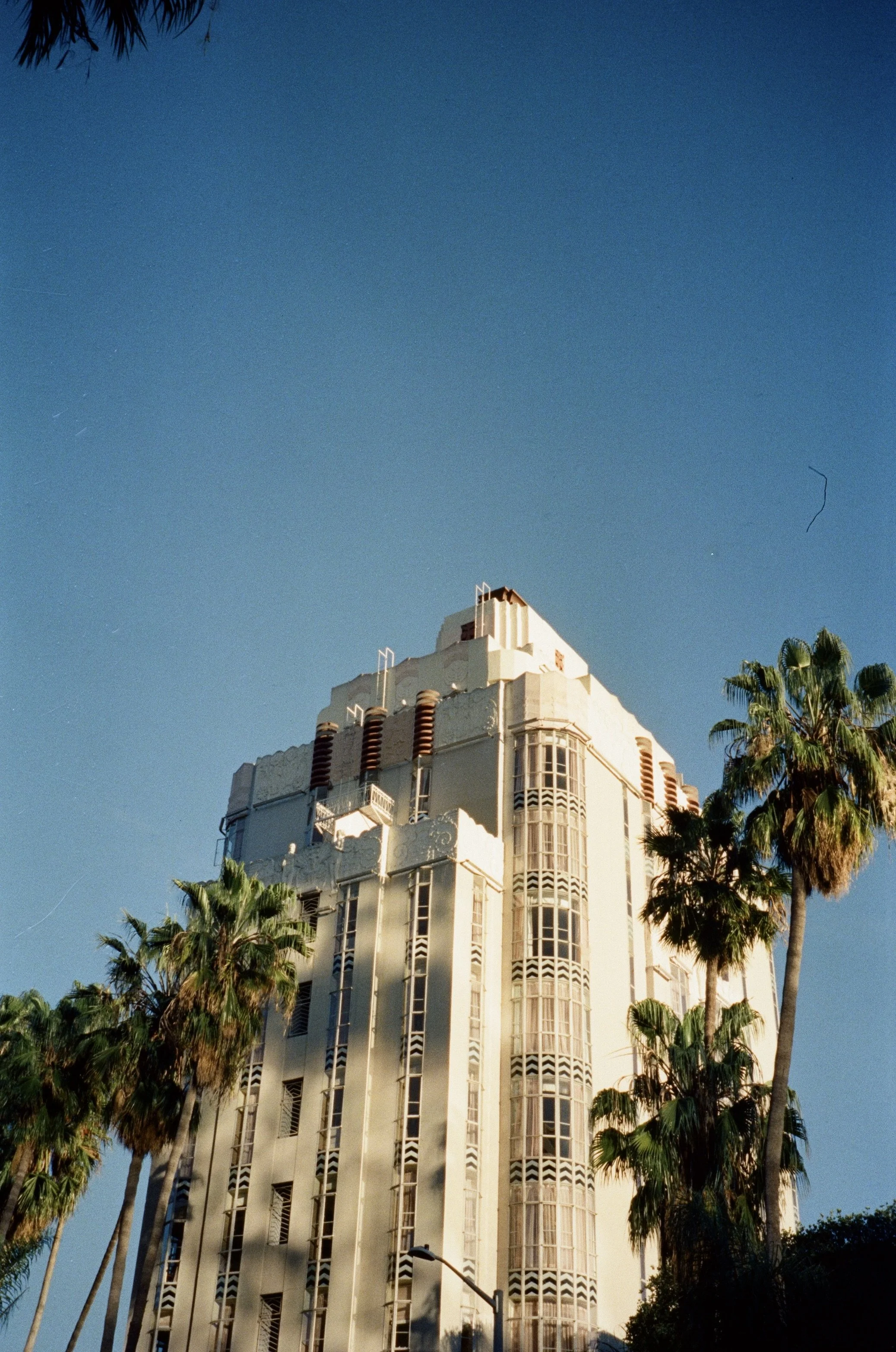 A white Art Deco building with vertical lines and curved windows, surrounded by tall palm trees, against a clear blue sky.