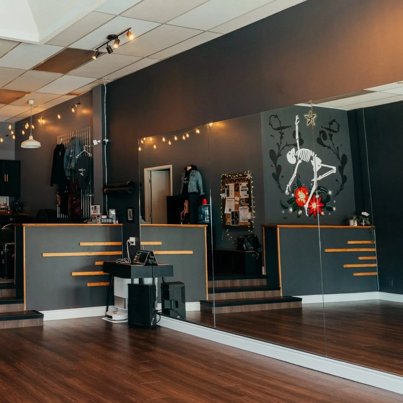An inside view of a dance or yoga studio with dark gray walls, a large mirror, wooden floor, and ceiling with track lighting and string lights. There are clothes hanging on the wall and various items on the counter, with stairs leading to a higher level decorated with wall art.