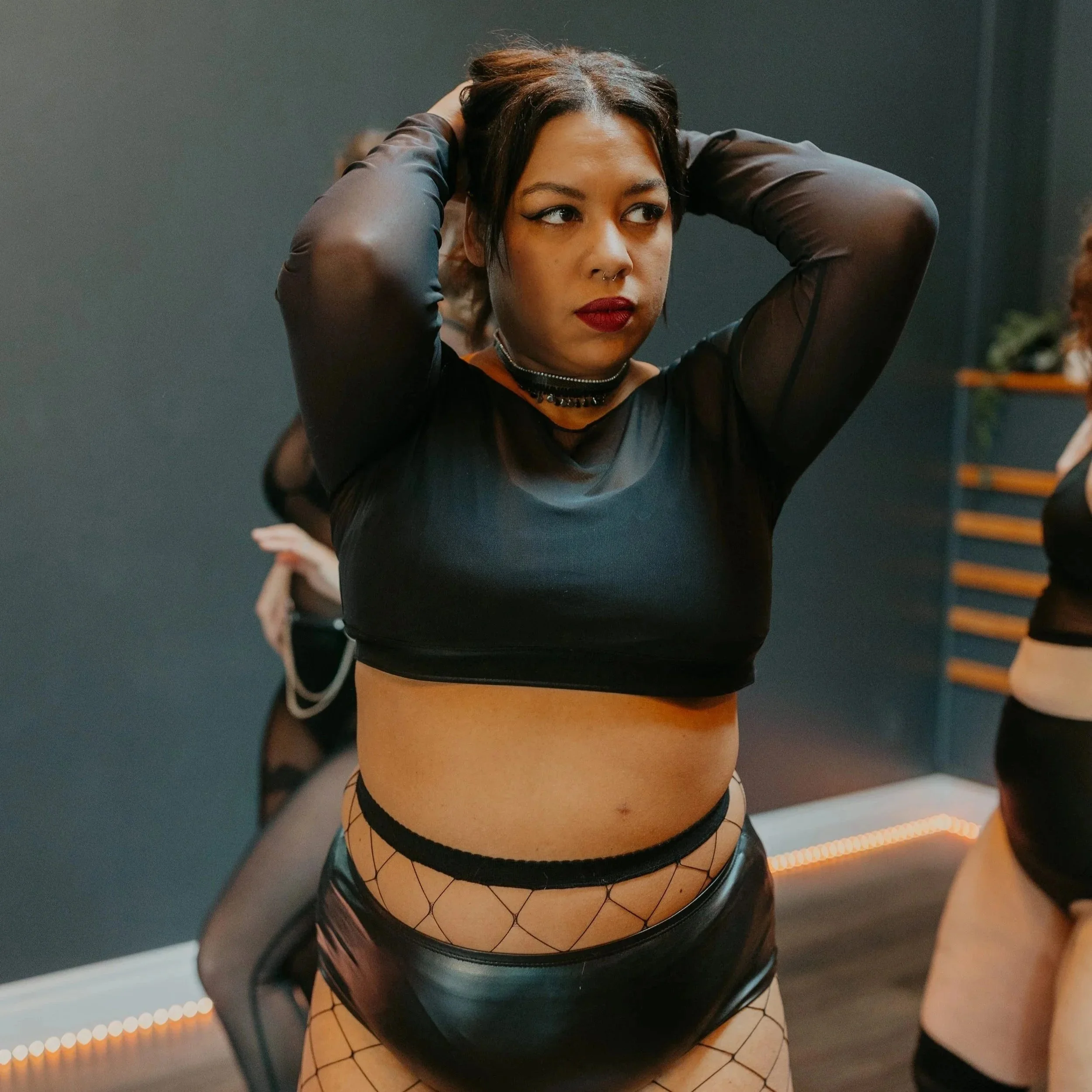 A woman of colous with dark hair and lipstick adjusting her hair in a black long-sleeve top, with black shorts, a choker necklace, posing with her hands in her hair in a dimly lit dance studio.
