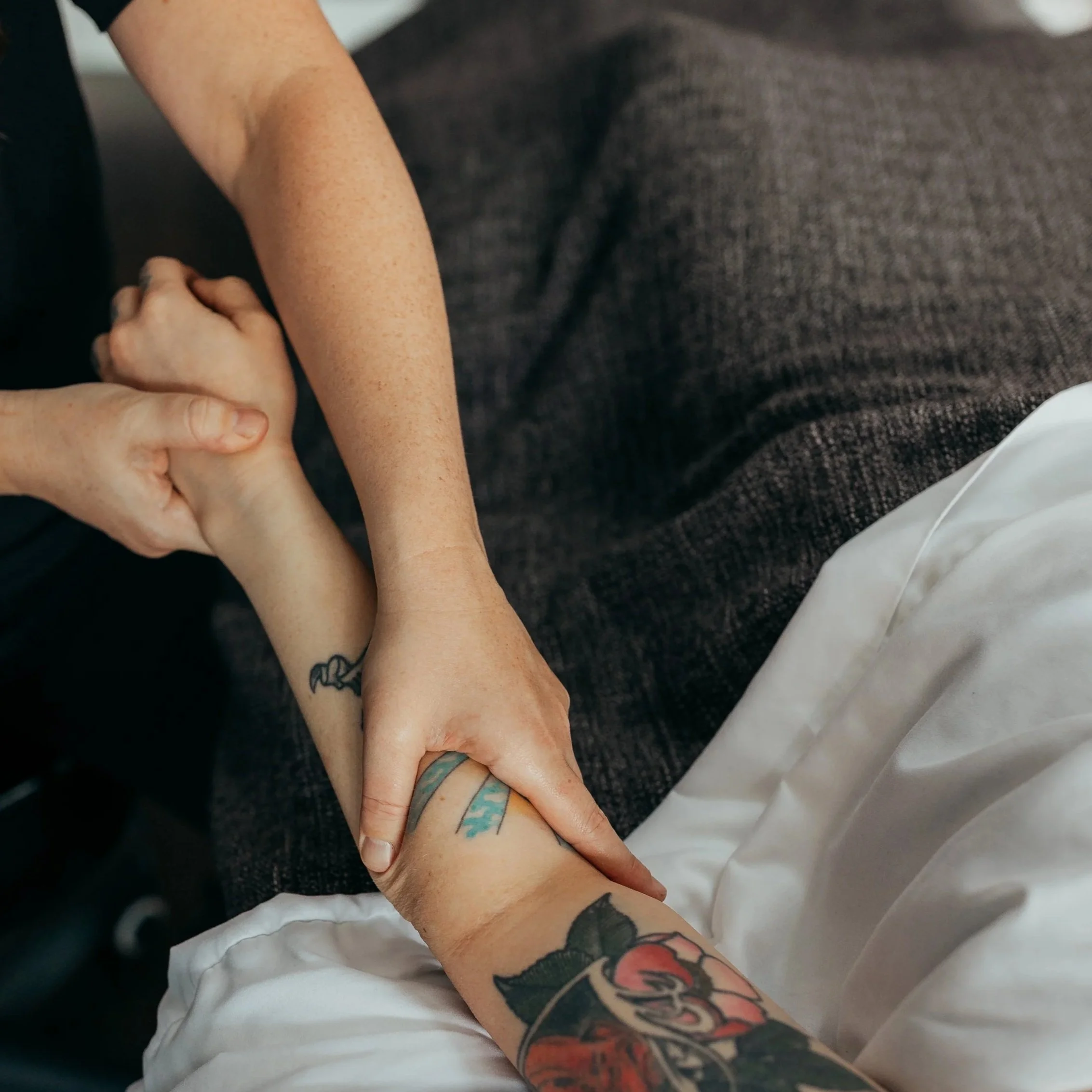 Close-up of a person performing massage therapy to another person's forearm and wrist, with tattoos visible on the arms, resting on a treatment table with clean white sheets.