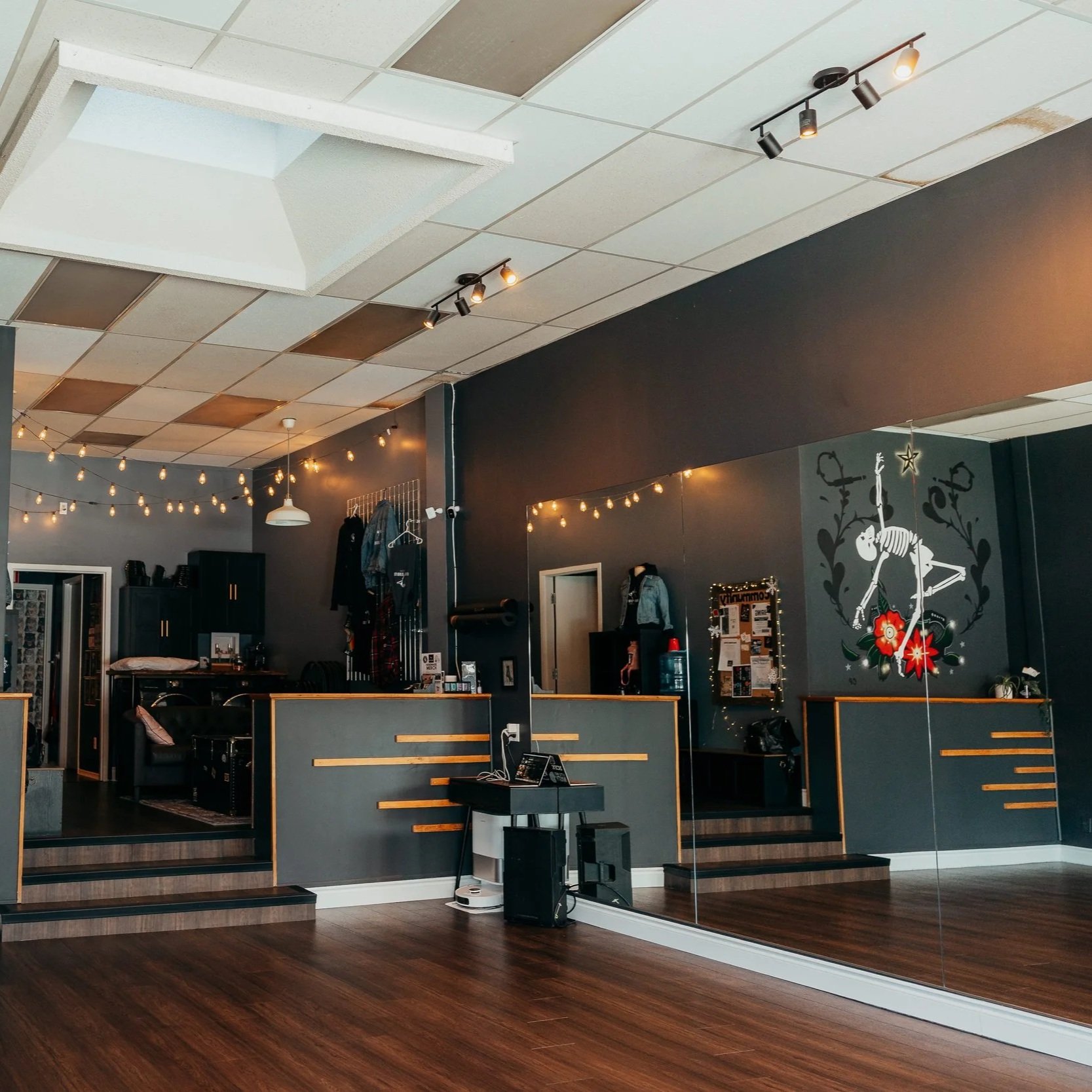 Interior of a dance studio with a large mirror on one wall, hardwood floors, dark walls with a skeleton and flowers mural, and string lights hanging from the ceiling.