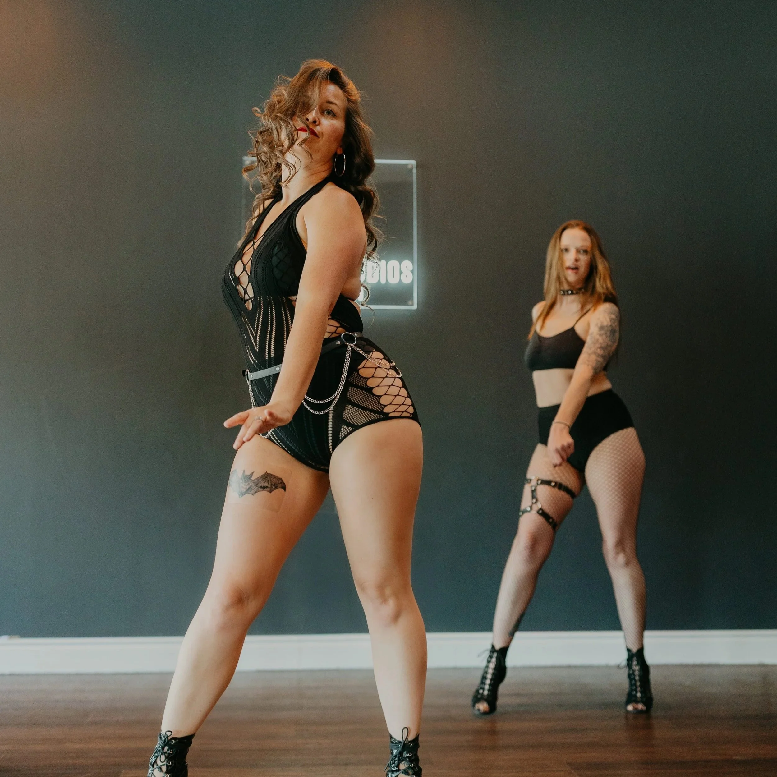 Two women in black dance costumes, one in the foreground and one in the background, performing a dance routine in a studio with a gray wall and wooden floor.