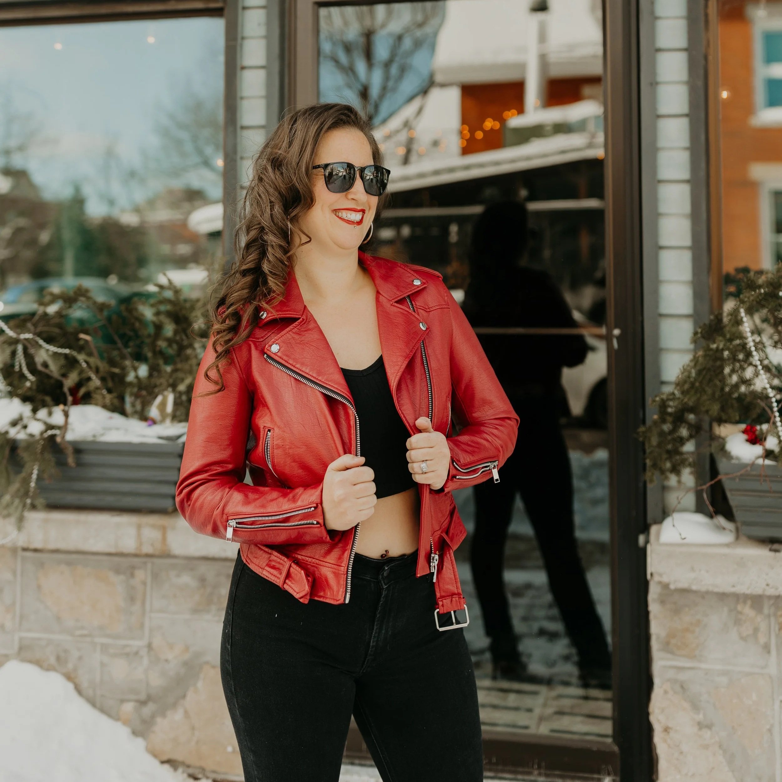 Woman with wavy brown hair wearing sunglasses, a red leather jacket, a black crop top, and black pants smiling and standing outside near a building with snow on the ground.