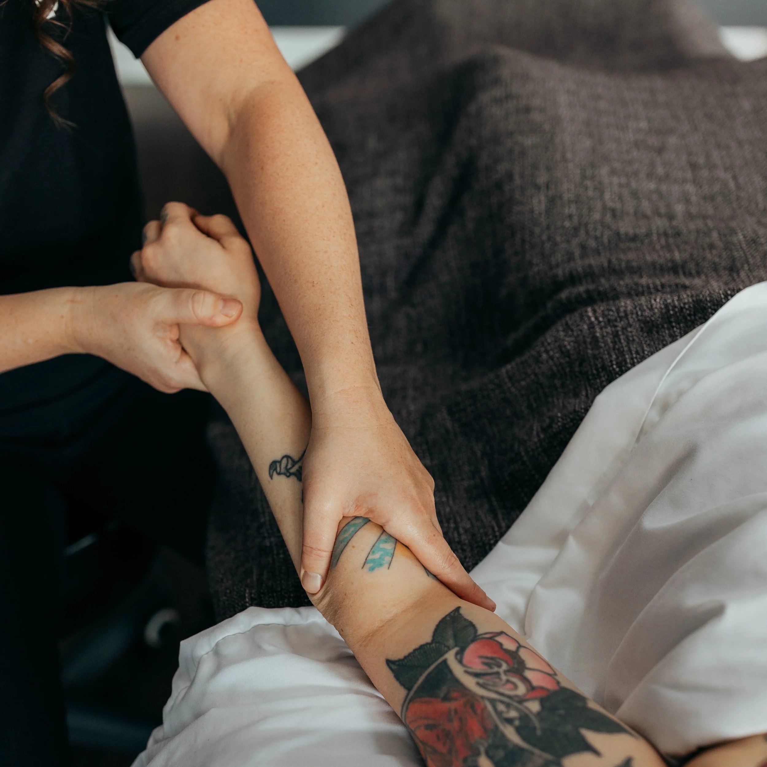 Close-up of a person performing massage therapy to another person's forearm and wrist, with tattoos visible on the arms, resting on a treatment table with clean white sheets.
