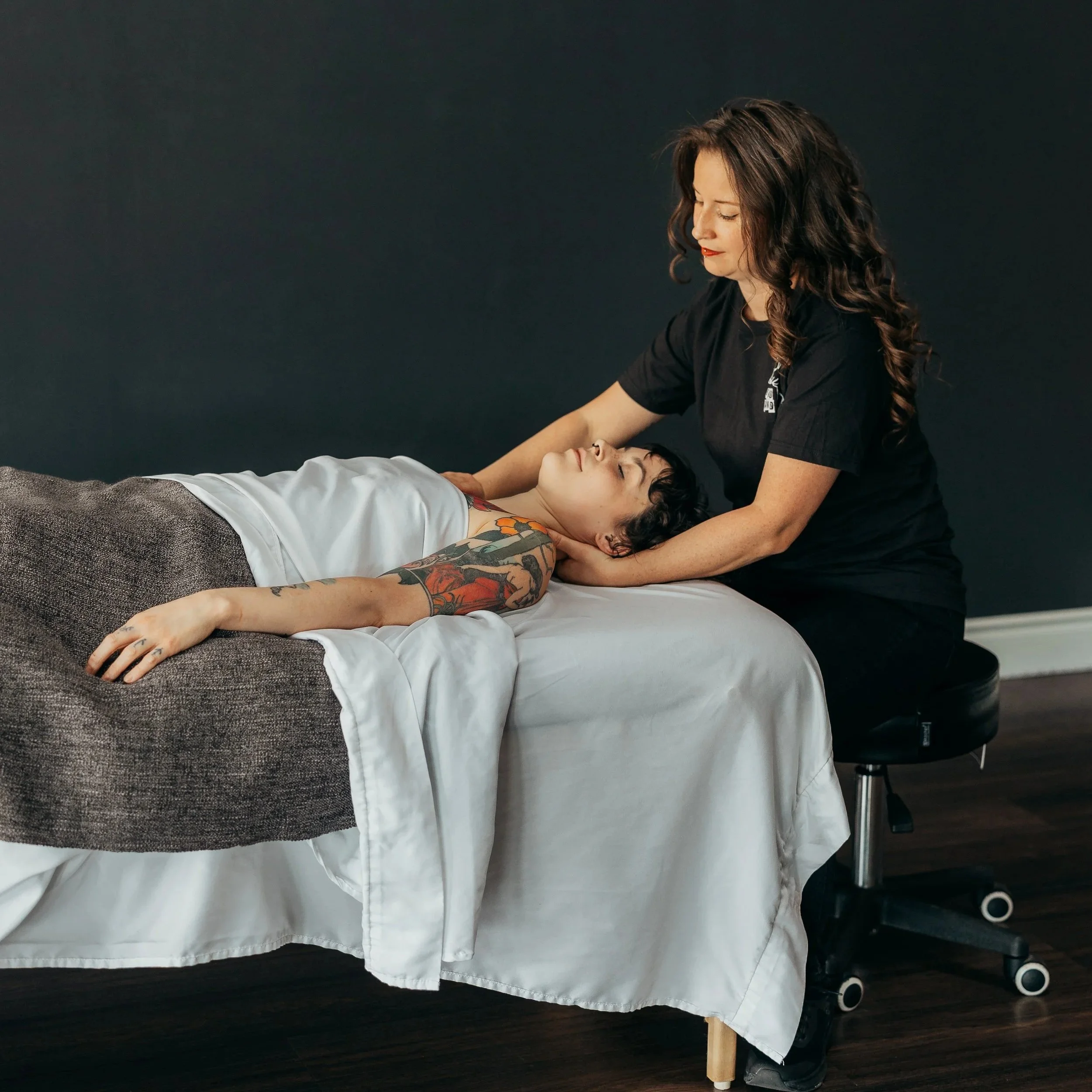 A tattooed woman receiving a neck massage from a massage therapist in a therapy room with a black wall background.